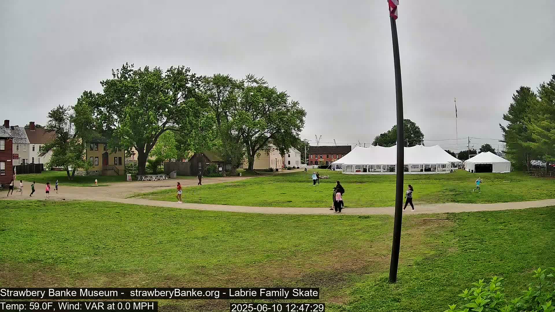 A grassy area with several people and children playing, historical-looking buildings, and a large white tent under an overcast sky.