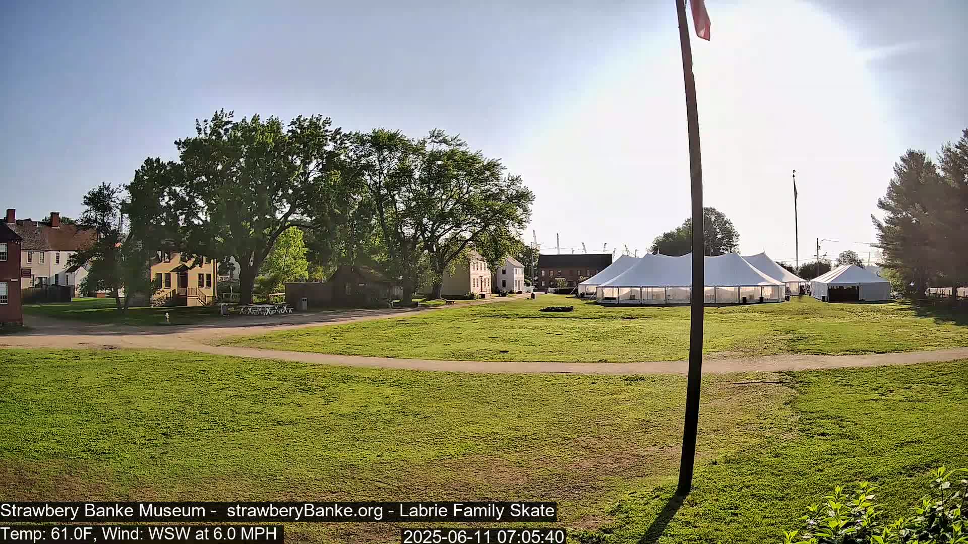 A sunny outdoor scene shows several historical-looking buildings and a large white tent in a grassy field, with a flagpole and American flag visible in the background.