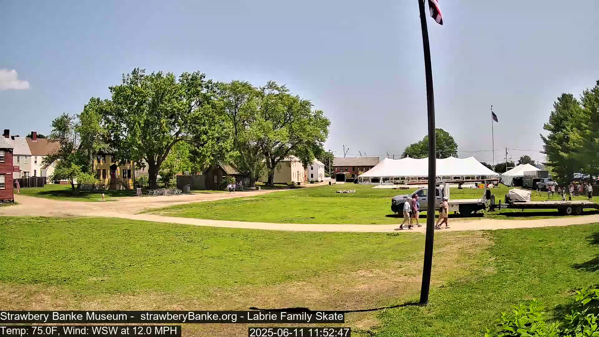 A sunny outdoor scene shows a grassy area with historical-looking buildings, a large white tent, a flatbed truck, and several people walking on a paved path.