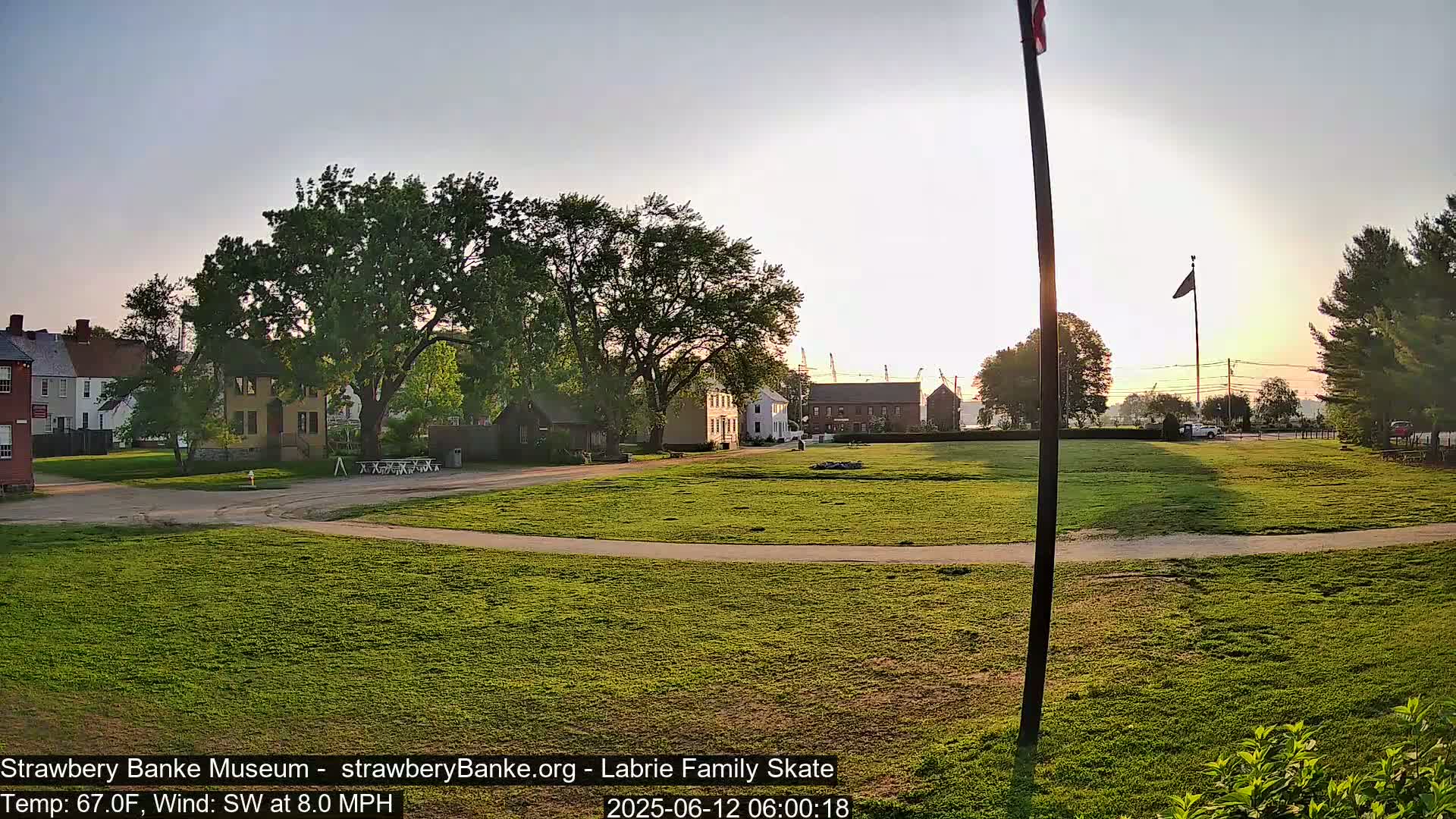 A grassy field with a few historical-looking buildings and trees in the background, under a clear, sunrise sky.