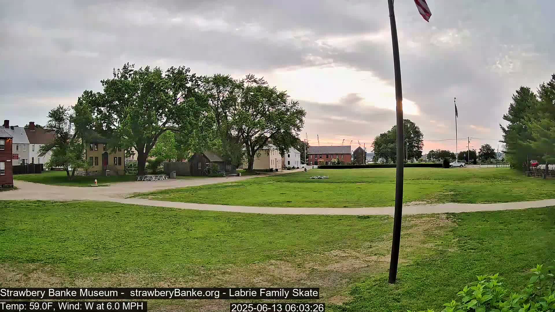 A grassy area with historical-looking buildings and trees under a partly cloudy sky at sunset.