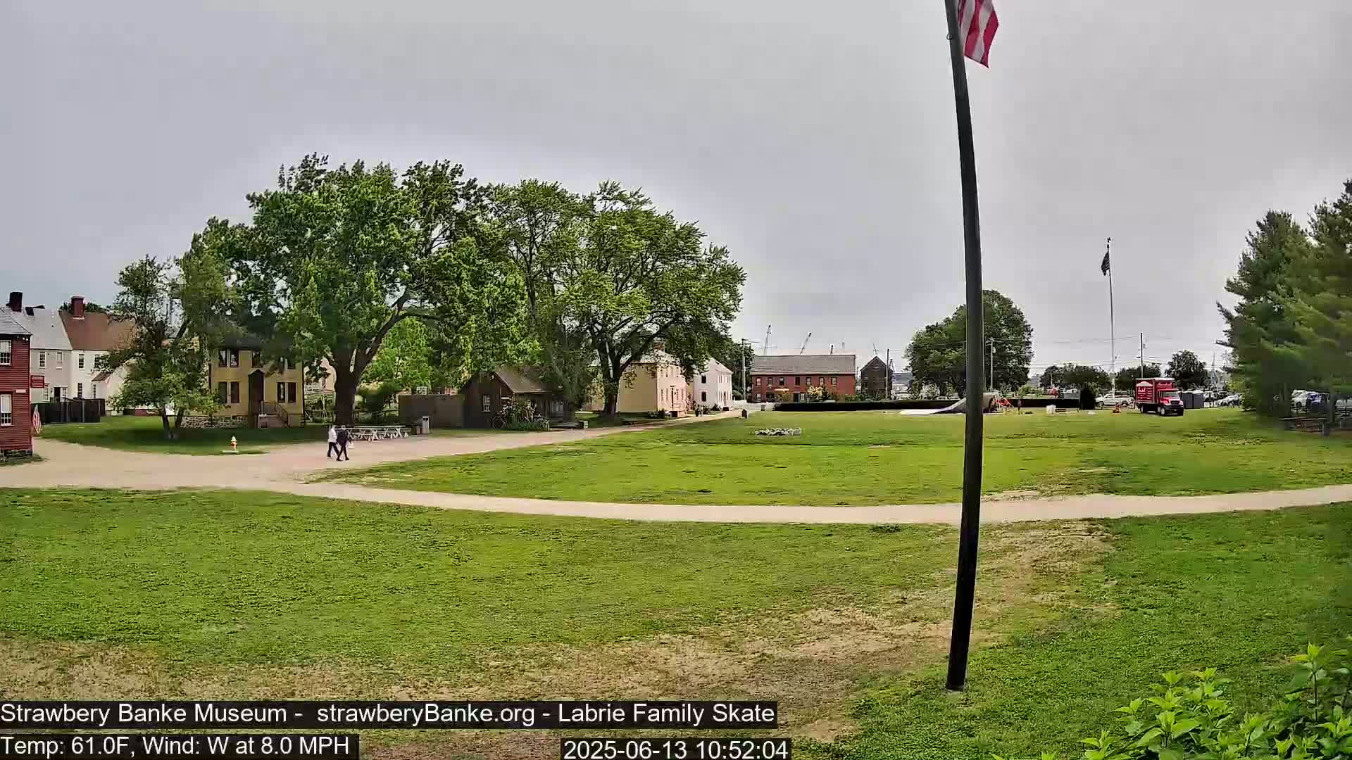 A grassy field with several historic-looking buildings and trees in the background under an overcast sky.