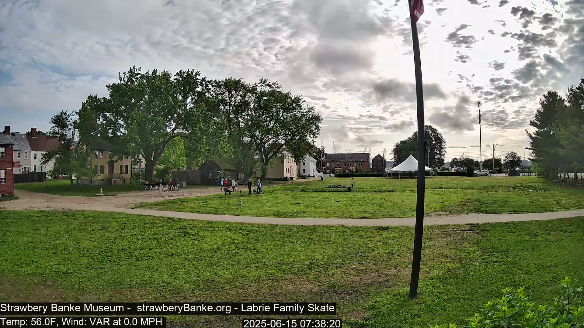 A grassy field with several historic buildings and a few people walking dogs under a partly cloudy sky.