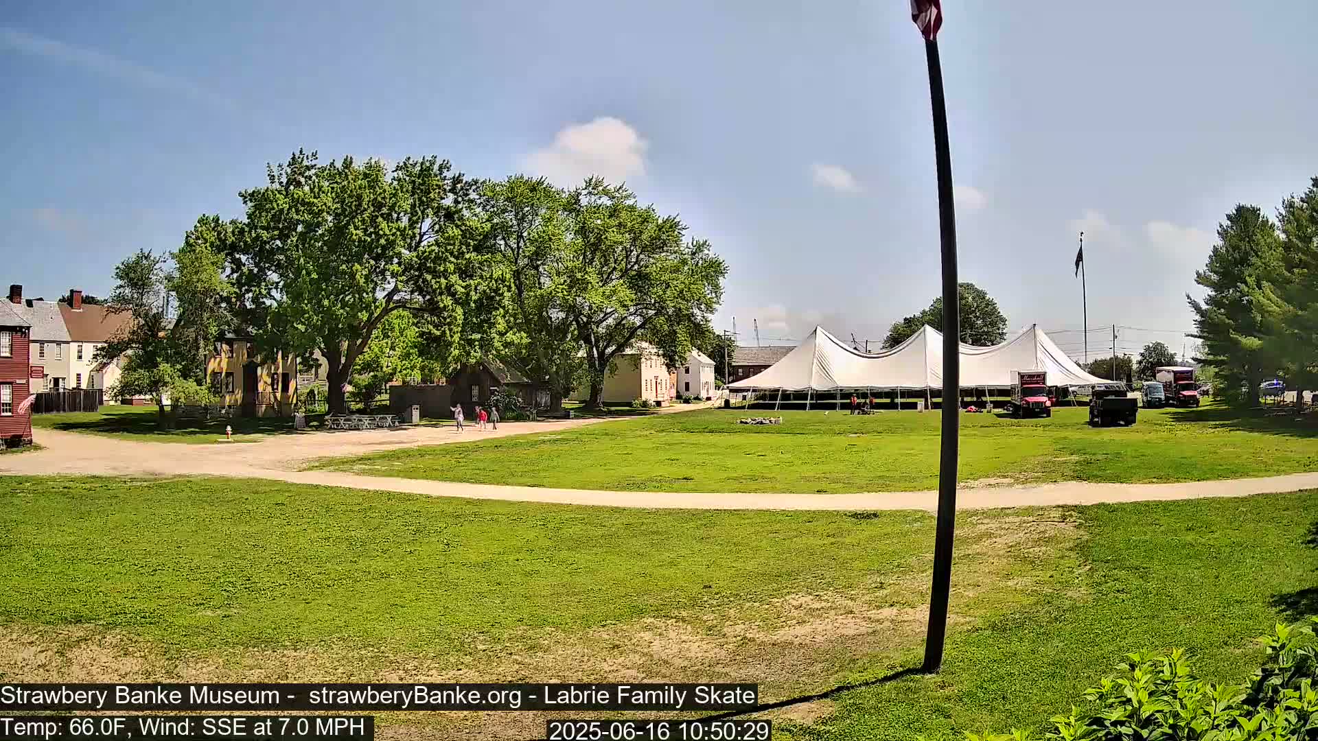 A large white tent sits on a grassy field in front of several historic-looking buildings under a sunny sky, with a light wind blowing.