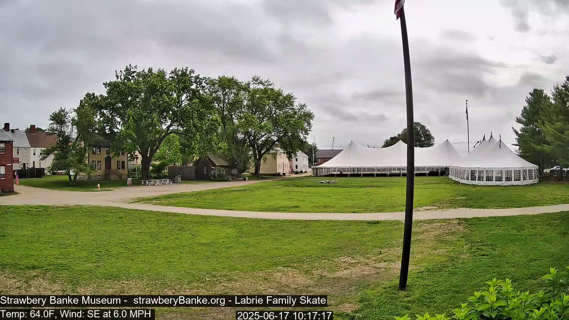 A large white tent sits on a grassy field next to several historic-looking buildings under an overcast sky.