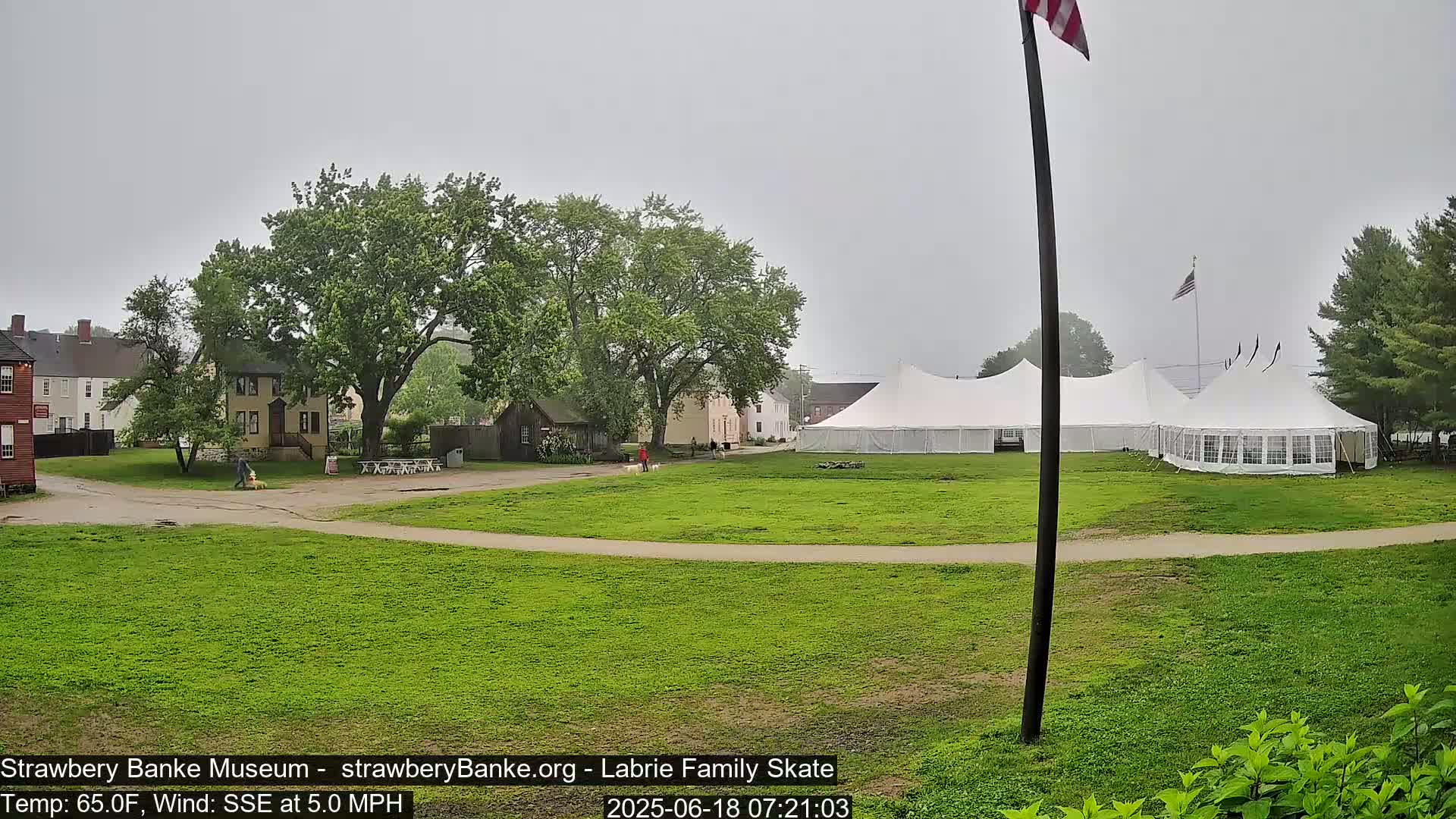 A large white tent sits on a grassy field in front of several historical-looking buildings on an overcast day.