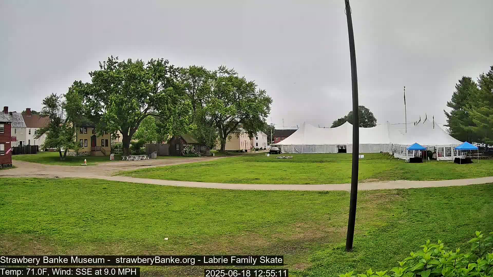 A grassy area with several historical-looking buildings, large white tents, and smaller blue tents under an overcast sky.