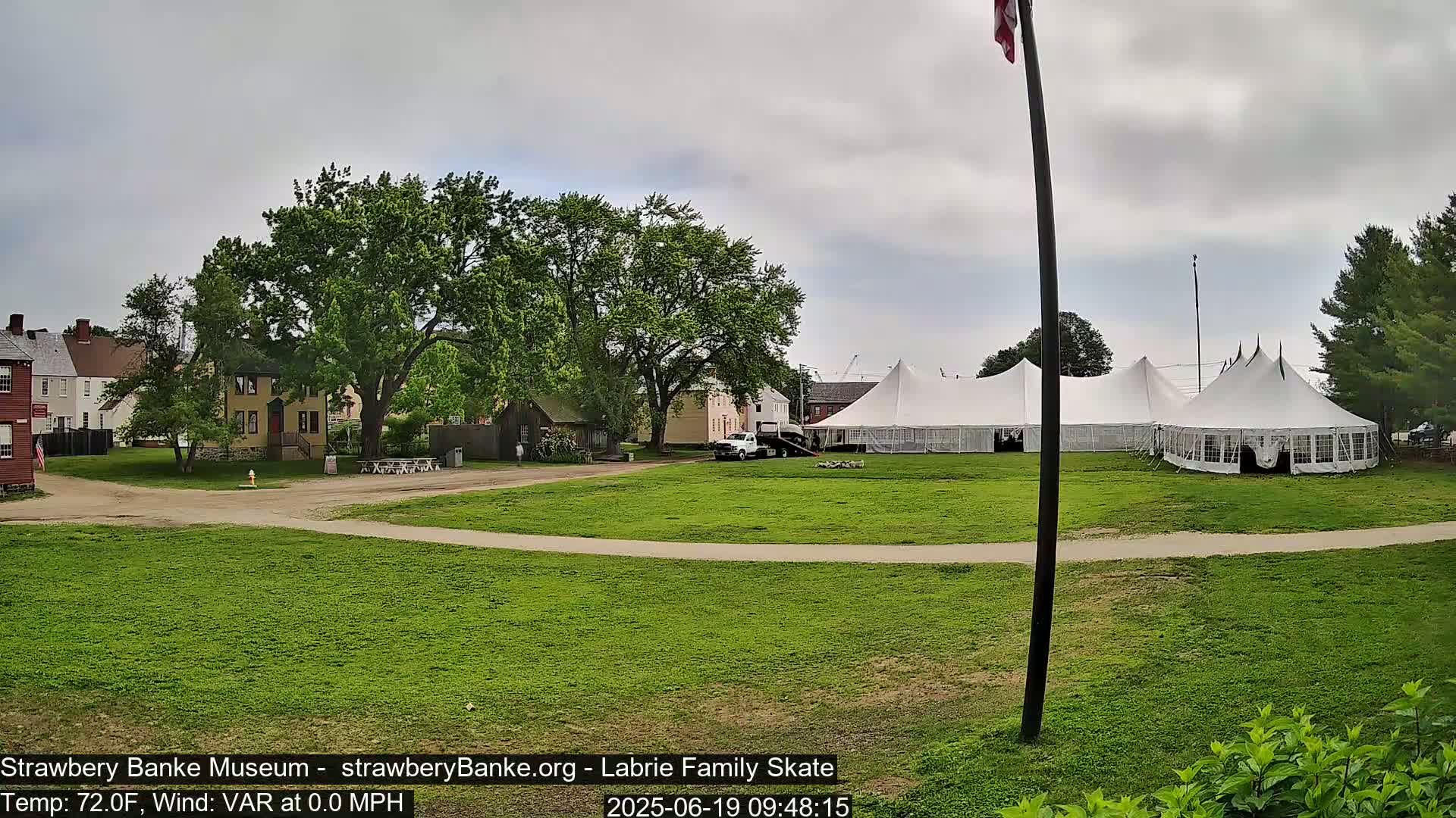 A grassy area with several historical-looking buildings, large white tents, and a partly cloudy sky.