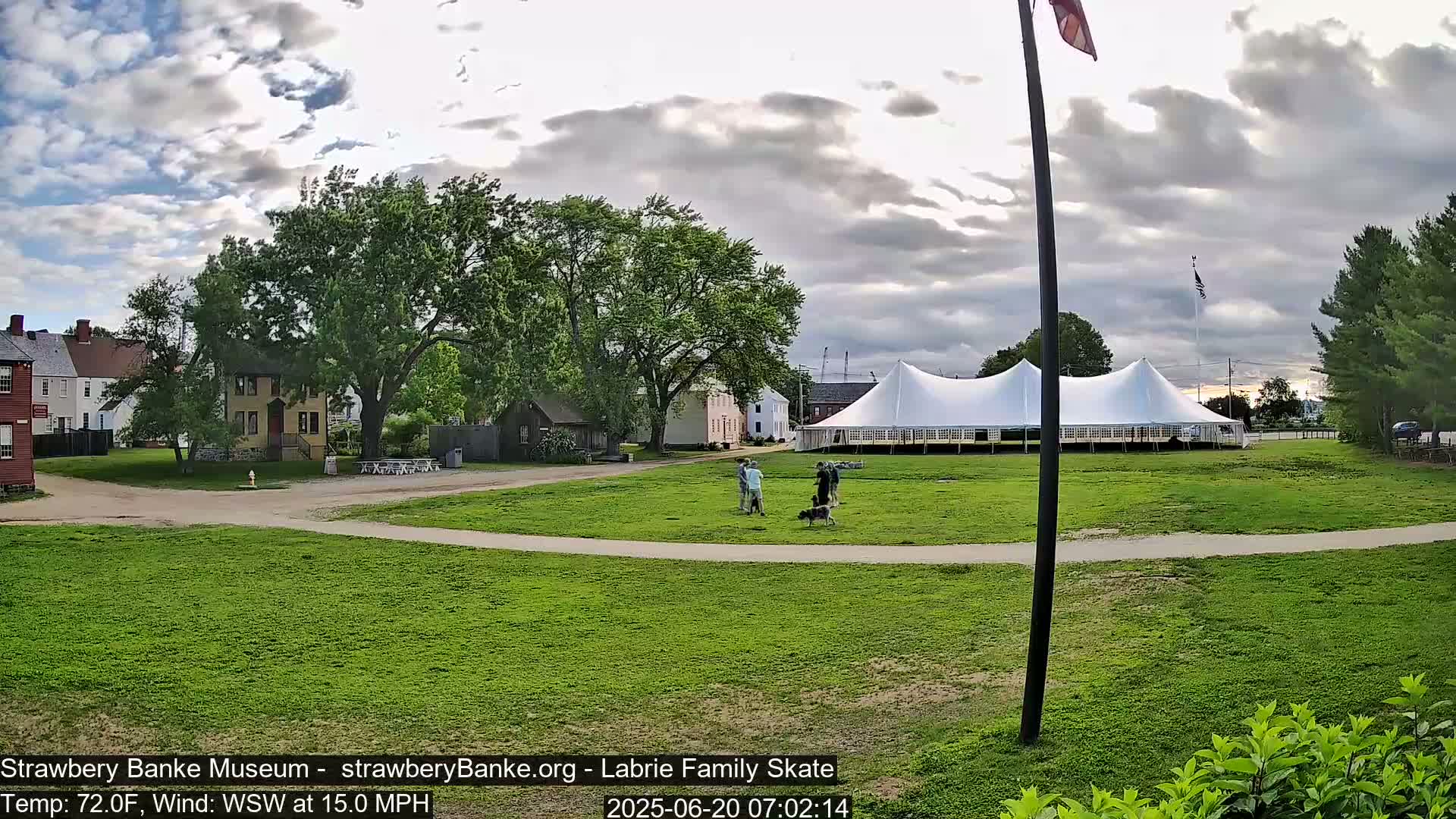 A large white tent sits on a grassy field next to several historical-looking buildings, under a partly cloudy sky, with a few people and dogs near the tent.