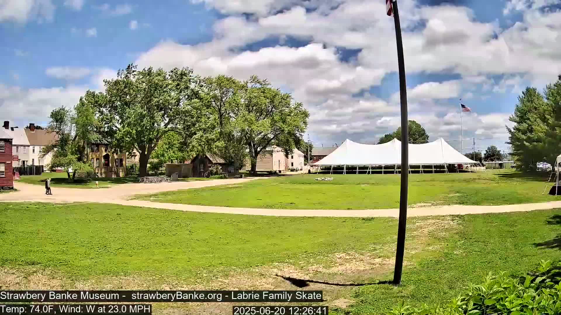 A large white tent sits on a grassy field next to several historic-looking buildings under a partly cloudy sky with a 23 mph west wind.