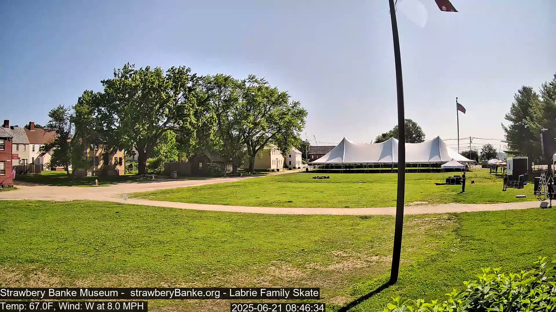 A large white tent sits on a grassy field in front of several historical-looking buildings under a clear, sunny sky.