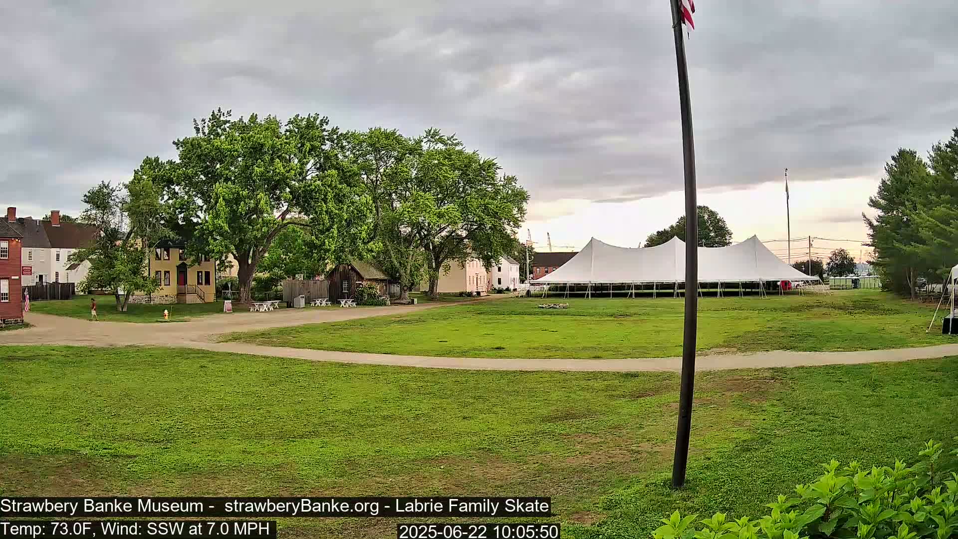 A large white tent sits on a grassy field in front of several historical-looking buildings under an overcast sky.