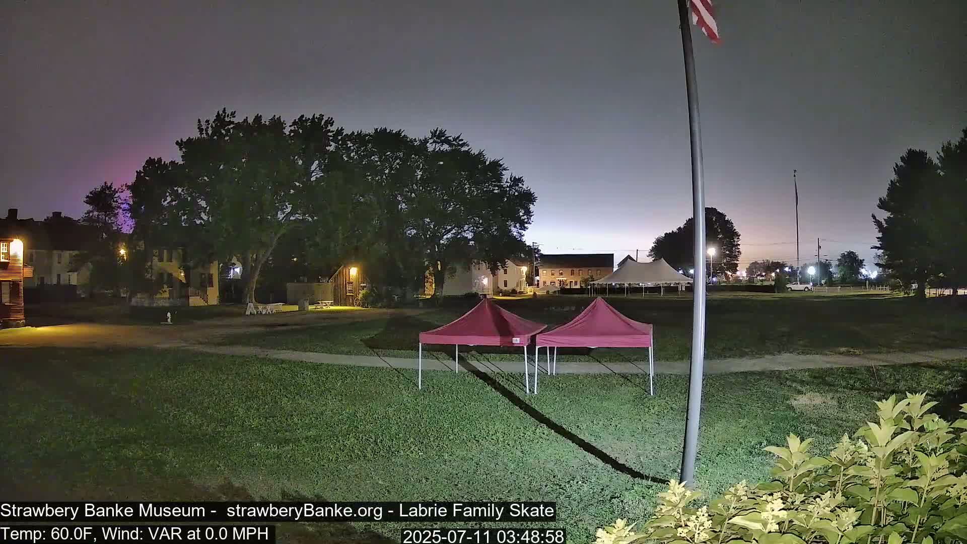 Two maroon pop-up tents sit on a grassy area at night, under a mostly clear, dusky sky, near some buildings and a larger tent in the background.