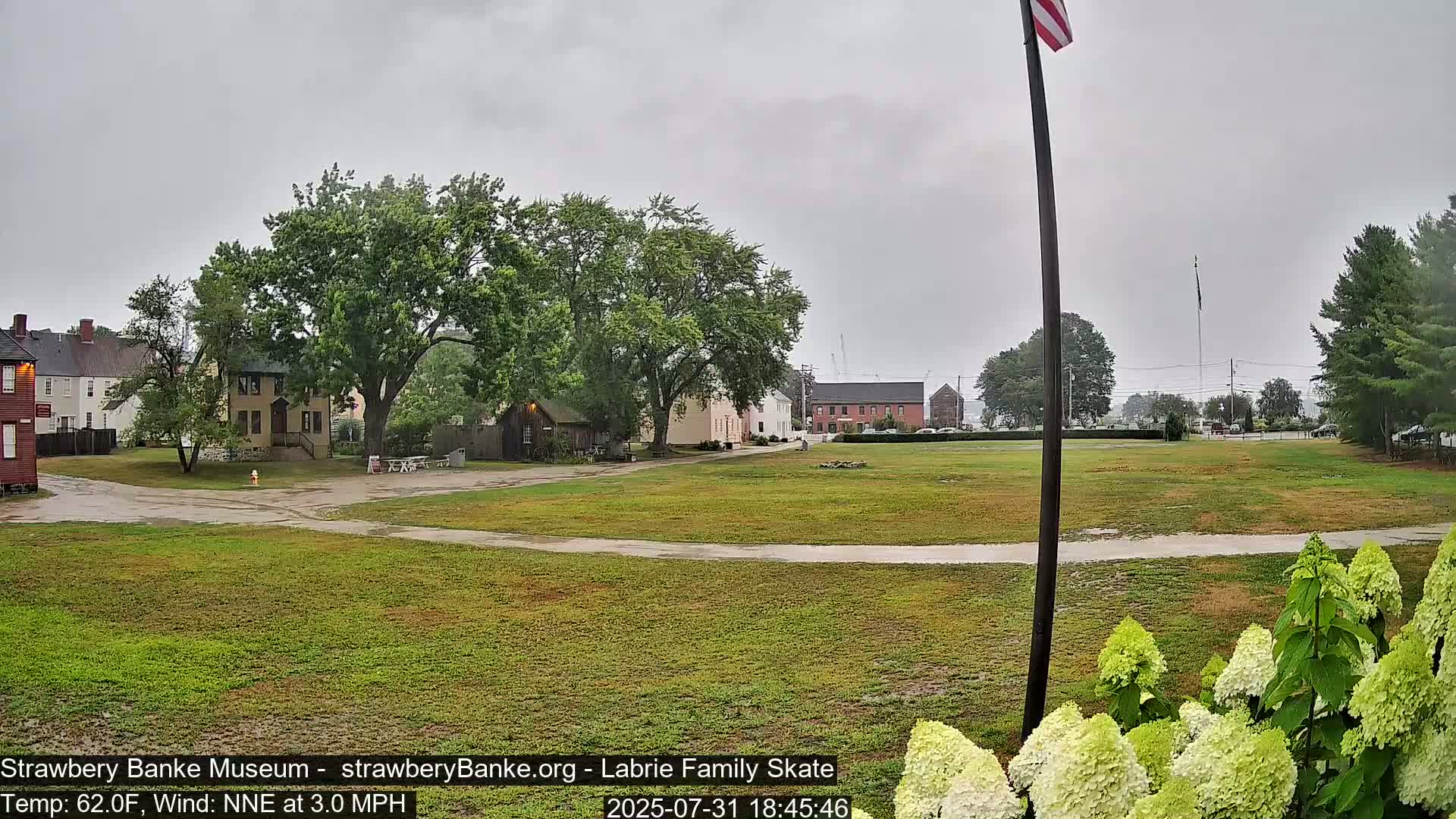 A grassy field with historical-looking buildings in the background under an overcast sky and light rain.