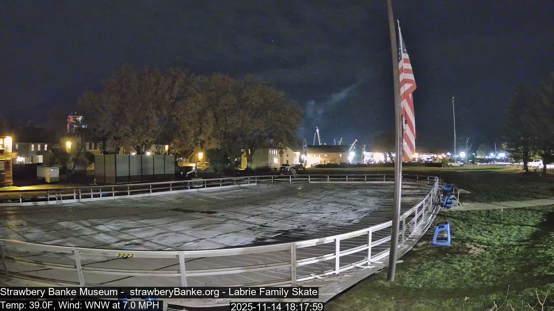 An empty, covered outdoor ice rink is visible at night under a clear sky, with an American flag standing nearby and distant illuminated buildings and trees forming the background.