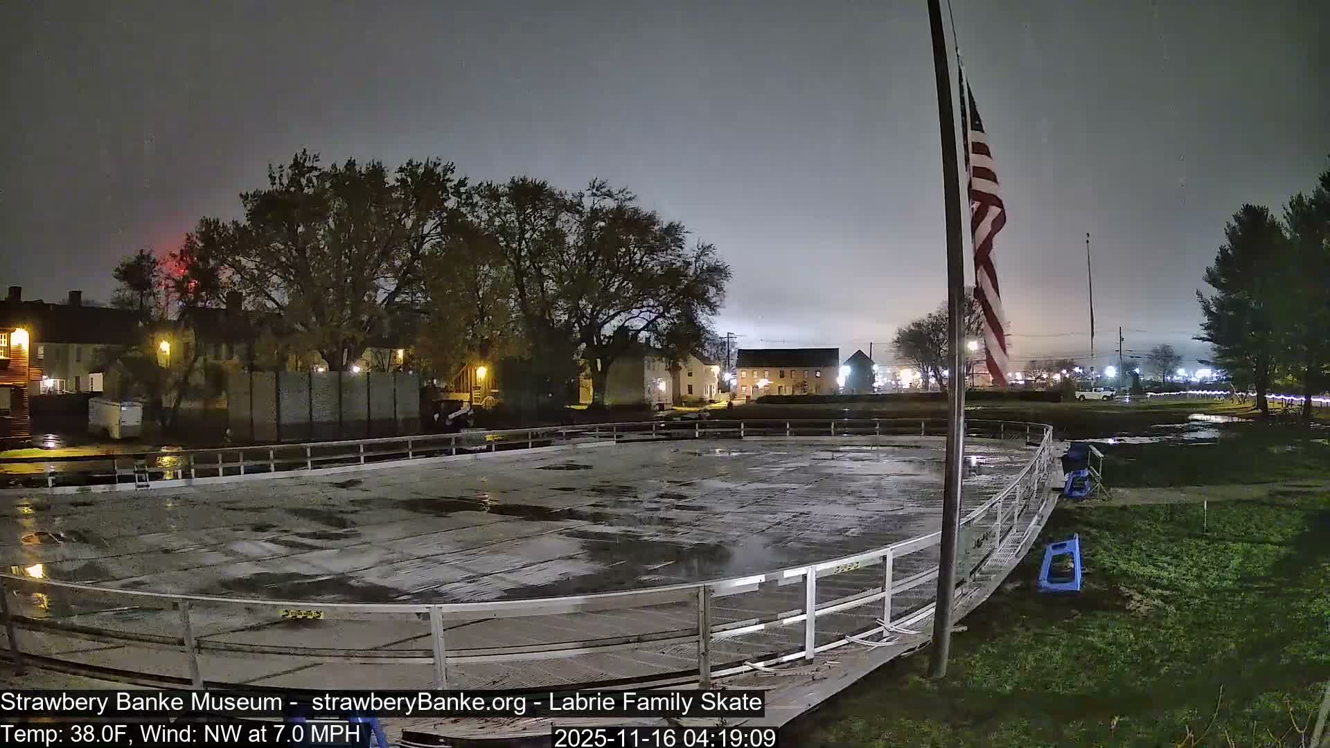 A wet, empty outdoor ice skating rink with visible puddles is illuminated at night under a dark, overcast sky, indicating recent or ongoing rain.