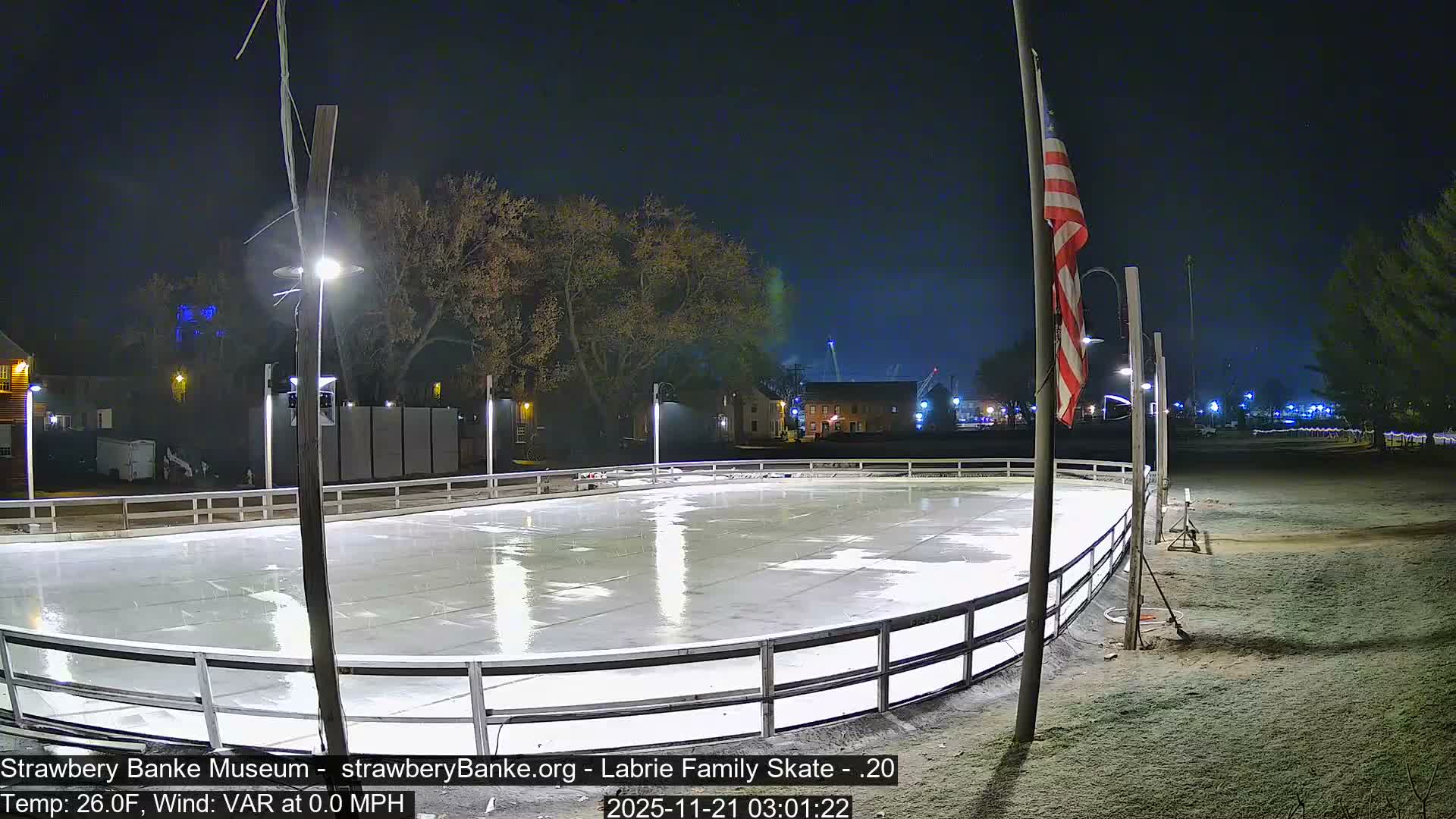 An empty outdoor ice rink is brightly illuminated at night, with an American flag on a pole and distant buildings visible under clear skies at 26.0F with no wind.