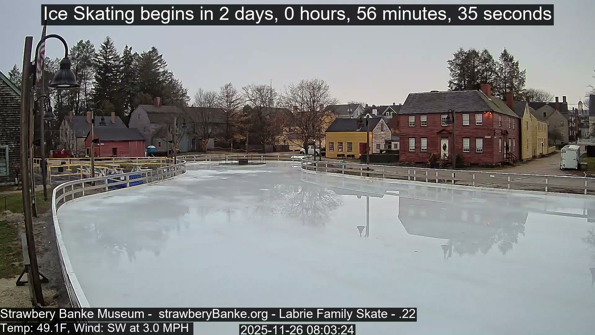 An empty, well-lit outdoor ice skating rink with a surrounding white barrier is visible at night, bordered by dormant trees and distant buildings, under a clear sky with a temperature of 33.1°F and a light WSW wind.