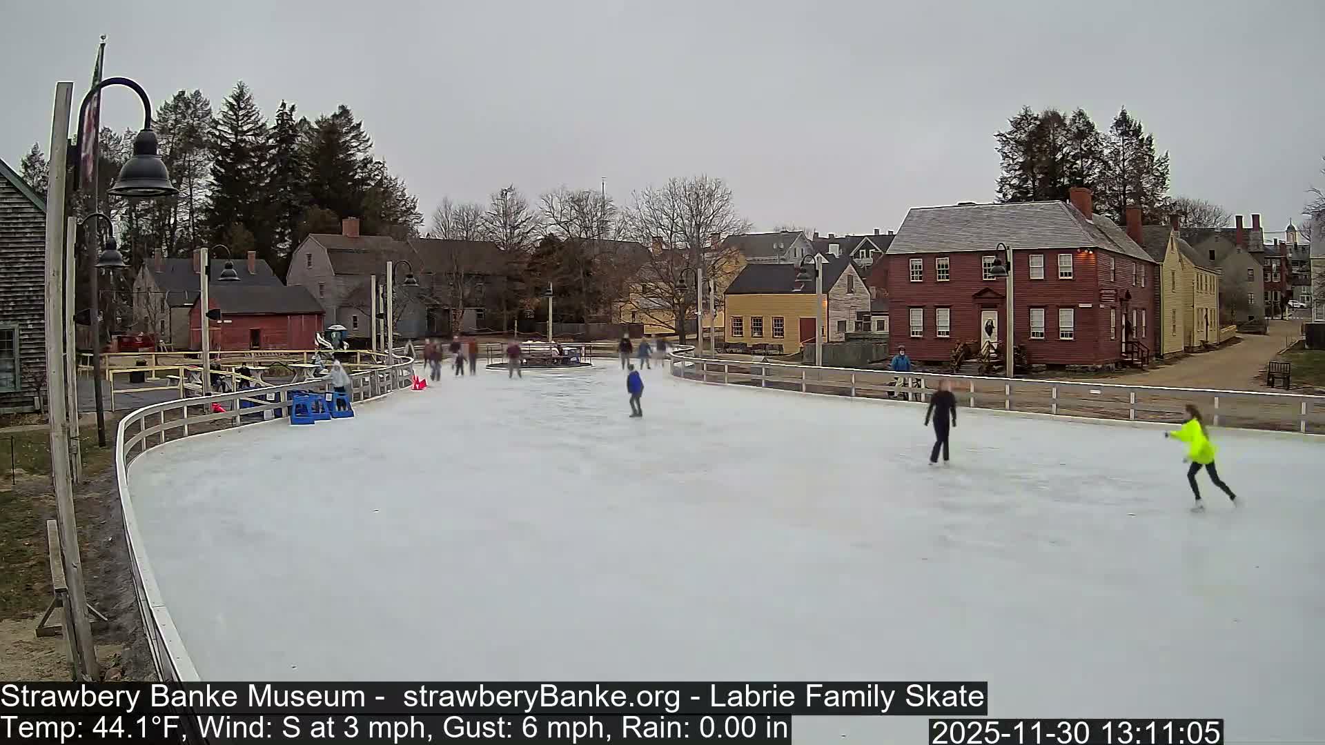 An empty, well-lit outdoor ice skating rink with a surrounding white barrier is visible at night, bordered by dormant trees and distant buildings, under a clear sky with a temperature of 33.1°F and a light WSW wind.