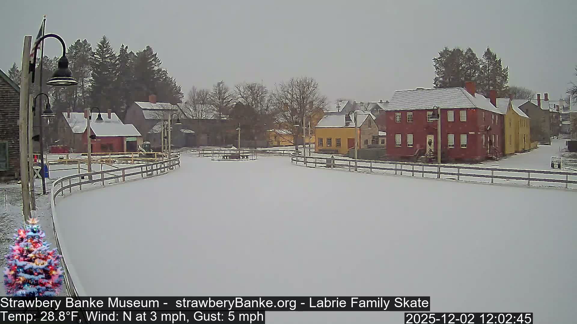 A historic village, featuring colorful buildings with snow-covered roofs, bare trees, and a wide snow-covered path or ice rink lined with railings, is depicted on an overcast winter day, with a brightly lit Christmas tree visible in the bottom left.