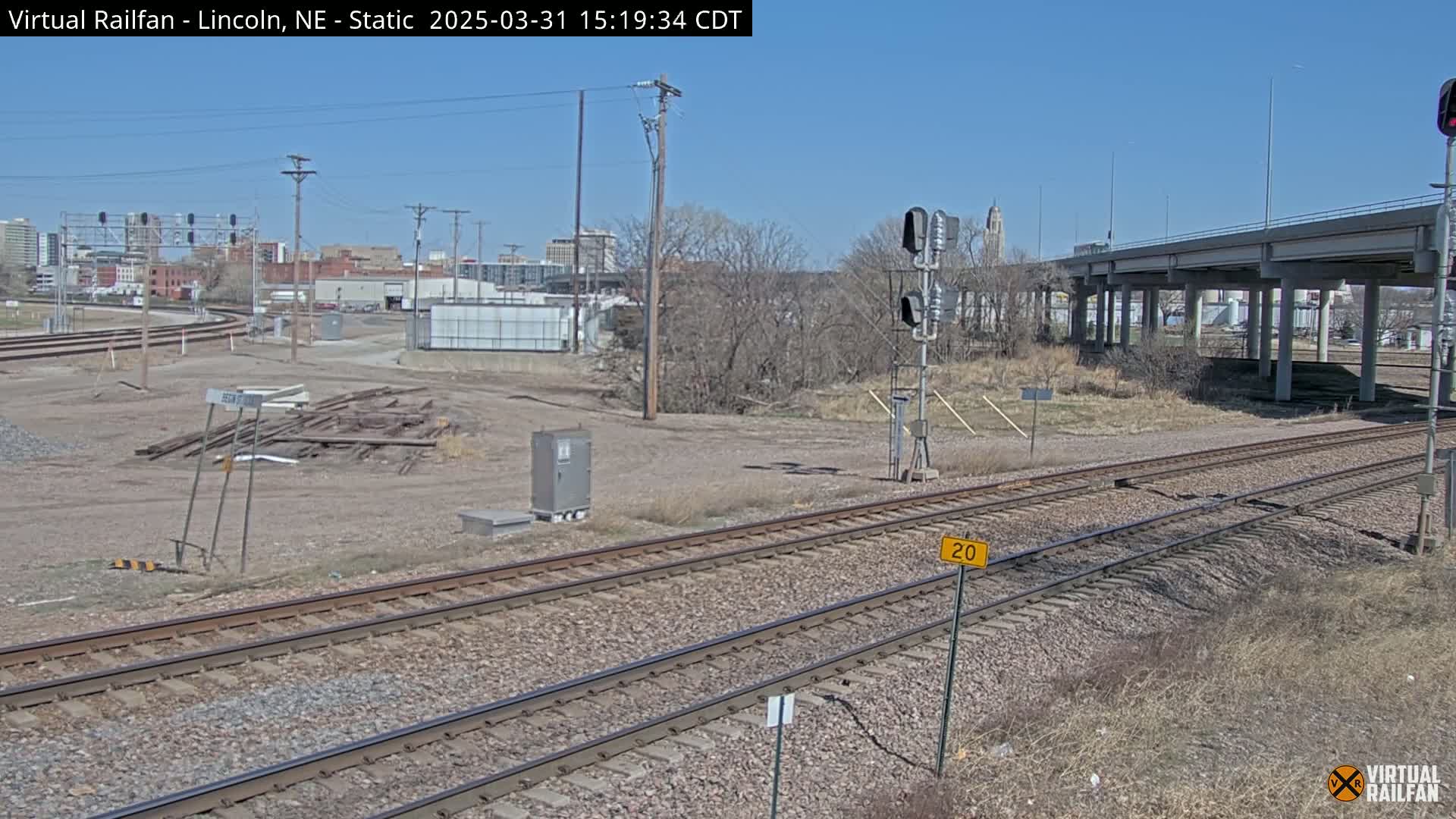 A view of railroad tracks on a clear, sunny day, with a highway overpass in the background and some bare trees and scrubby vegetation nearby.