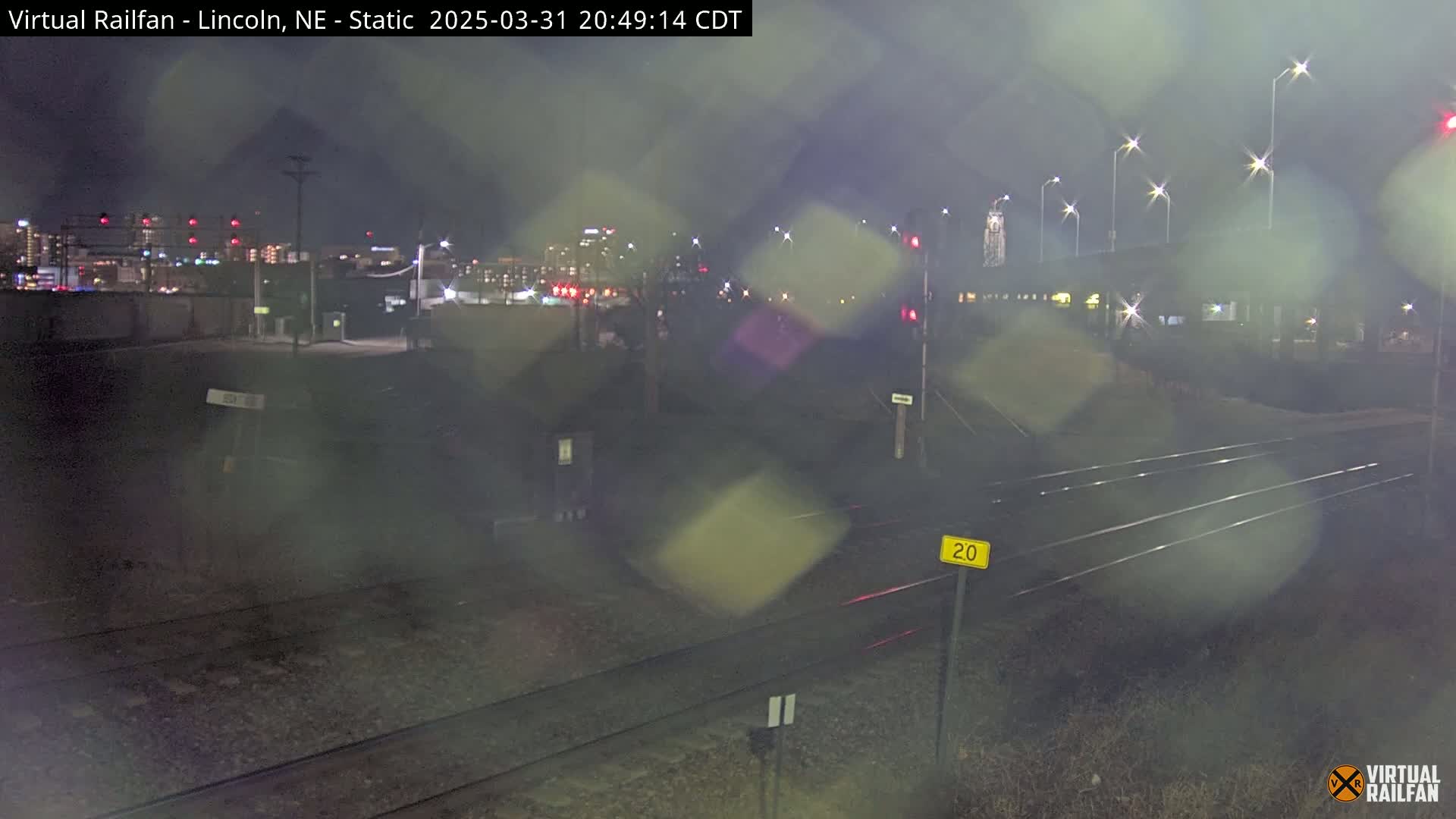 A nighttime view of railroad tracks and a city skyline in the distance, under rainy conditions.