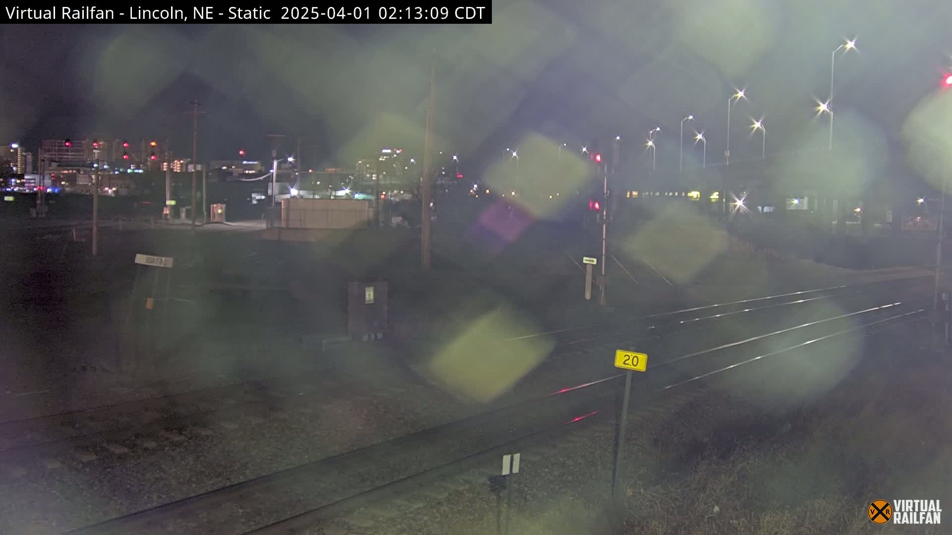 A nighttime view of railroad tracks and a city skyline under a rainy sky.