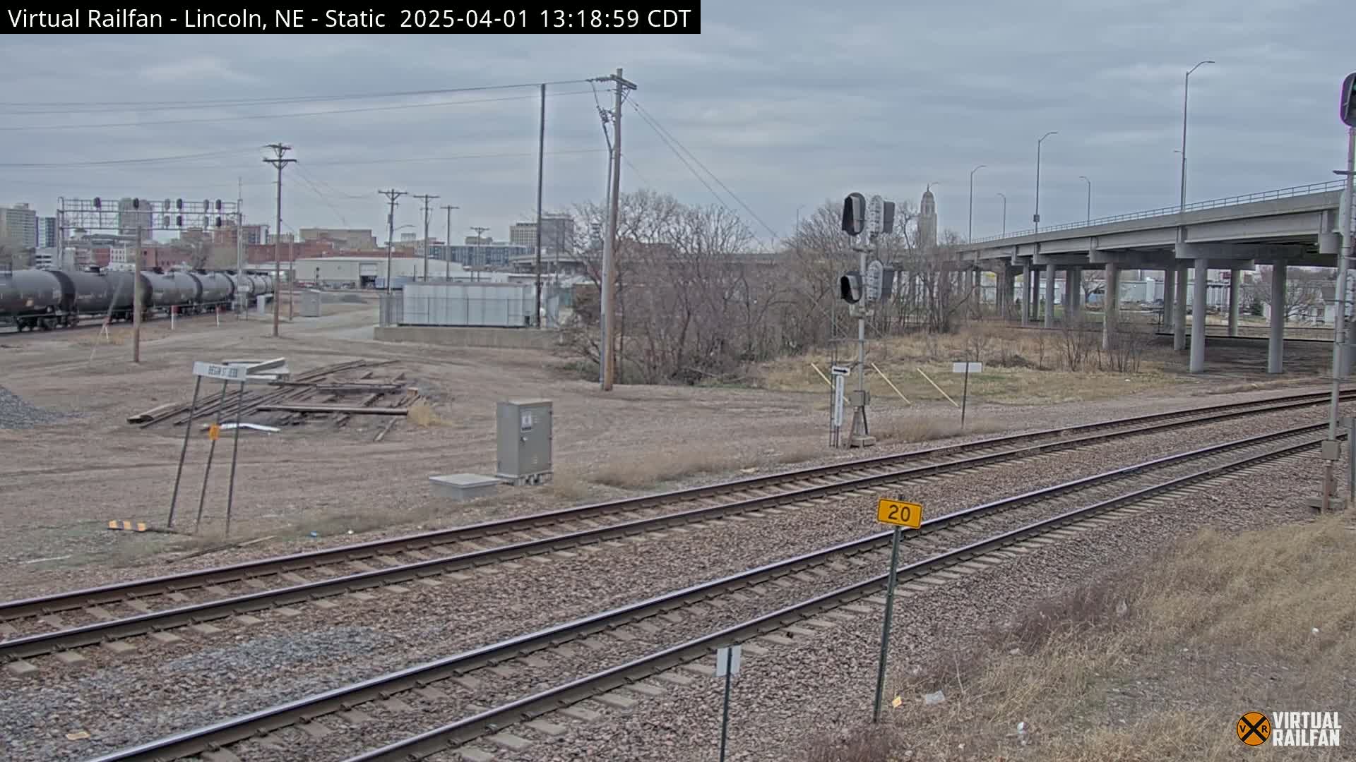 A train track with a freight train in the distance under an overcast sky, passing beneath a large overpass.