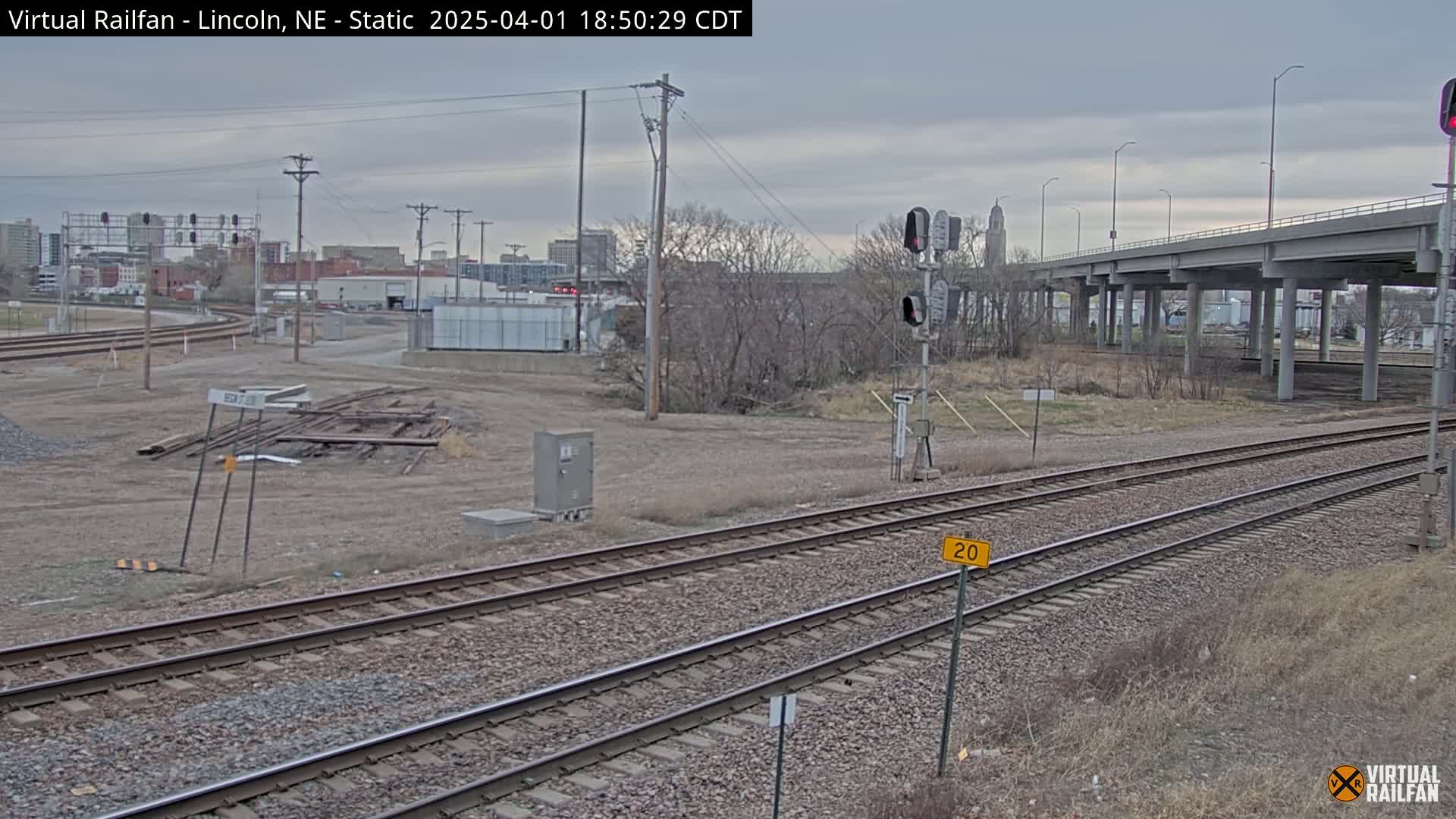 A view of railroad tracks under an overpass on an overcast day, with a city skyline in the background.