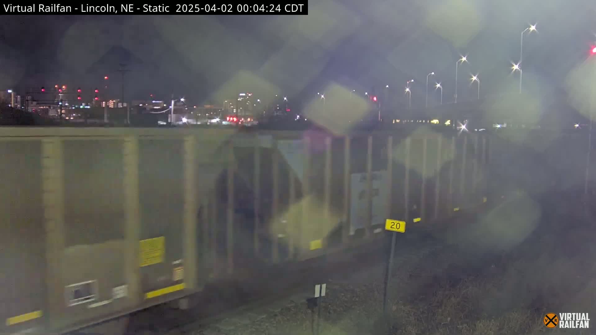 A long freight train moves through a city at night in rainy conditions, passing under streetlights.