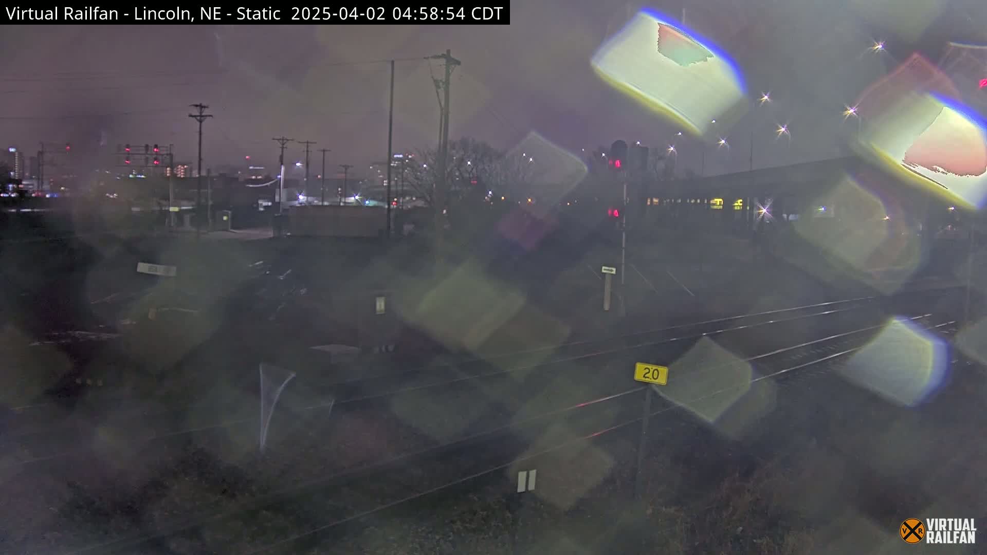 A nighttime view of railroad tracks and a city skyline in rainy conditions, with blurry lights and reflections obscuring some details.