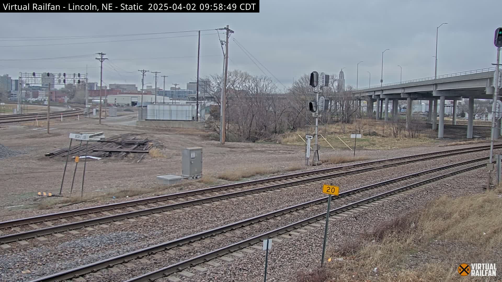 Multiple sets of train tracks run alongside a highway overpass under an overcast sky, with bare trees and industrial buildings in the background.