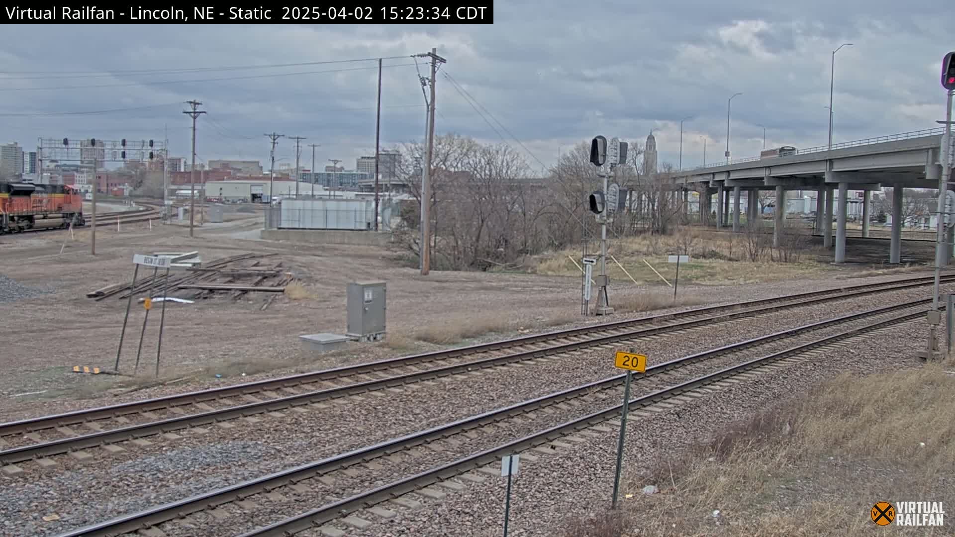 Under a cloudy sky, a view of railroad tracks stretches towards a distant cityscape with a freeway overpass in the background, and a diesel locomotive is parked to the left.