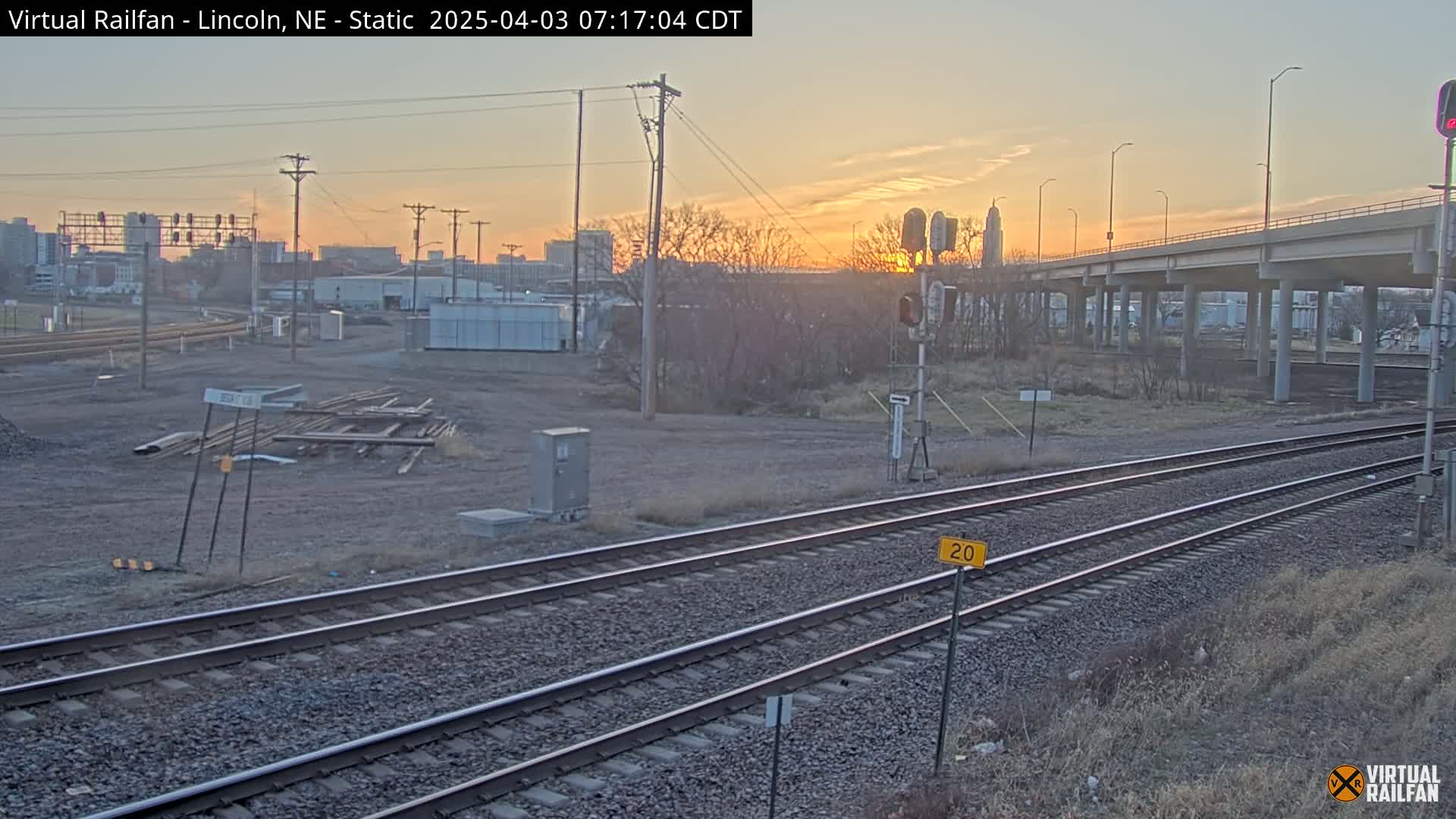 A sunrise illuminates a cityscape behind railroad tracks and a highway overpass, with bare winter trees and a construction site in the foreground.