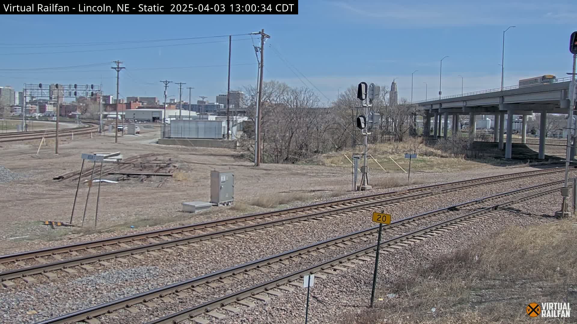 A view of train tracks under a clear sky, with a city skyline and a highway overpass in the background.