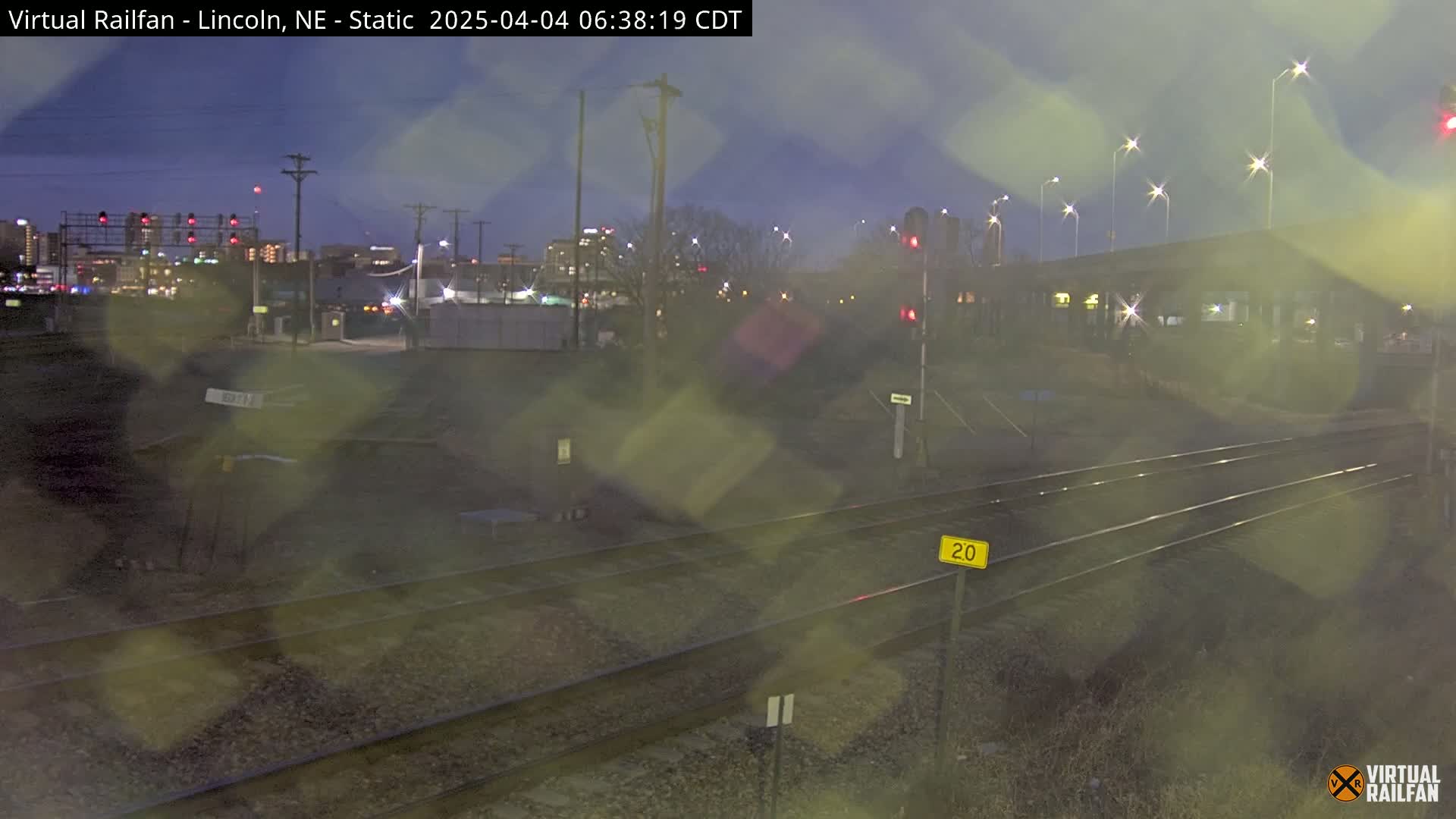 A nighttime view of railroad tracks in the rain, with city lights and buildings visible in the distance.
