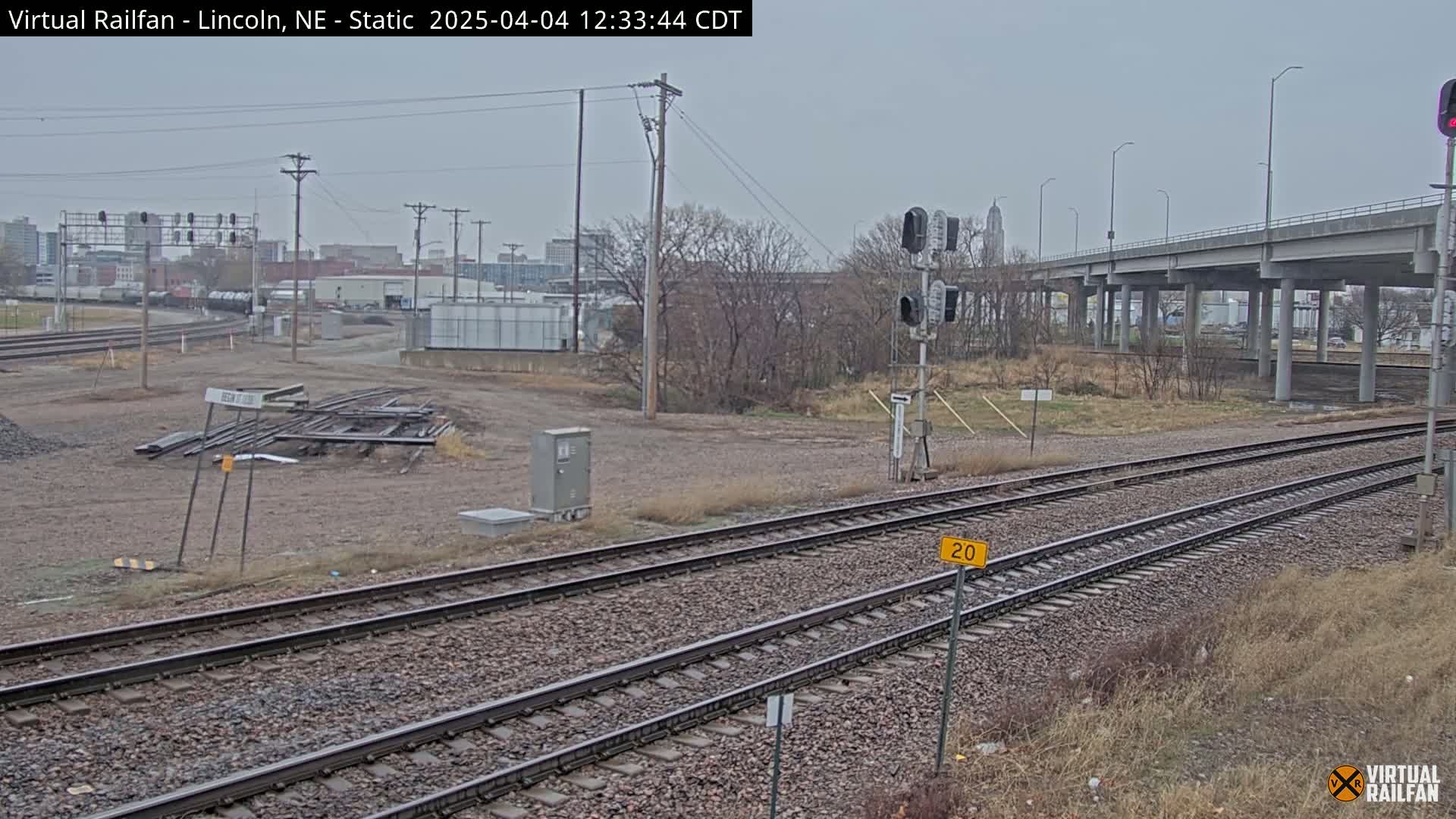 A view of railroad tracks on an overcast day, with a highway overpass in the background and bare trees lining the sides.