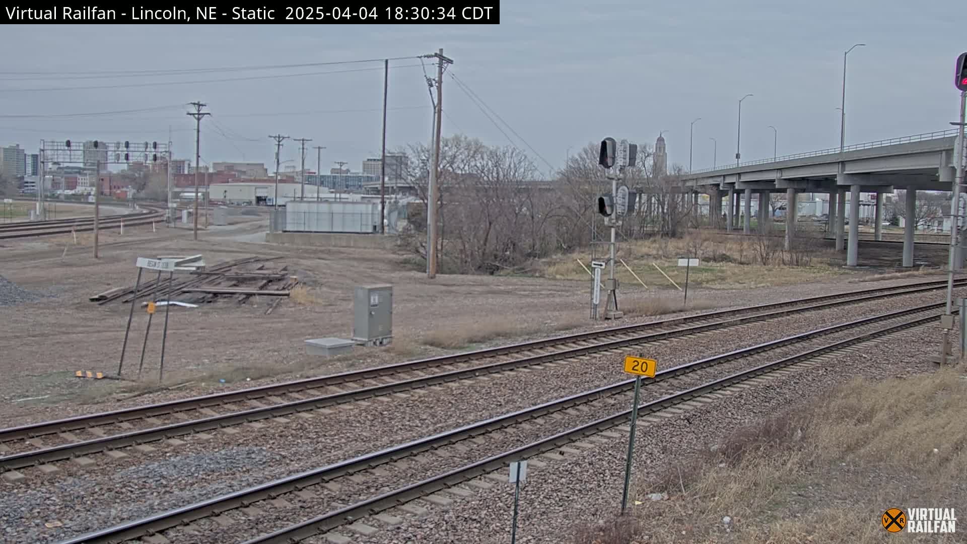 A view of railroad tracks under an overpass on an overcast day, with a city skyline in the distance.