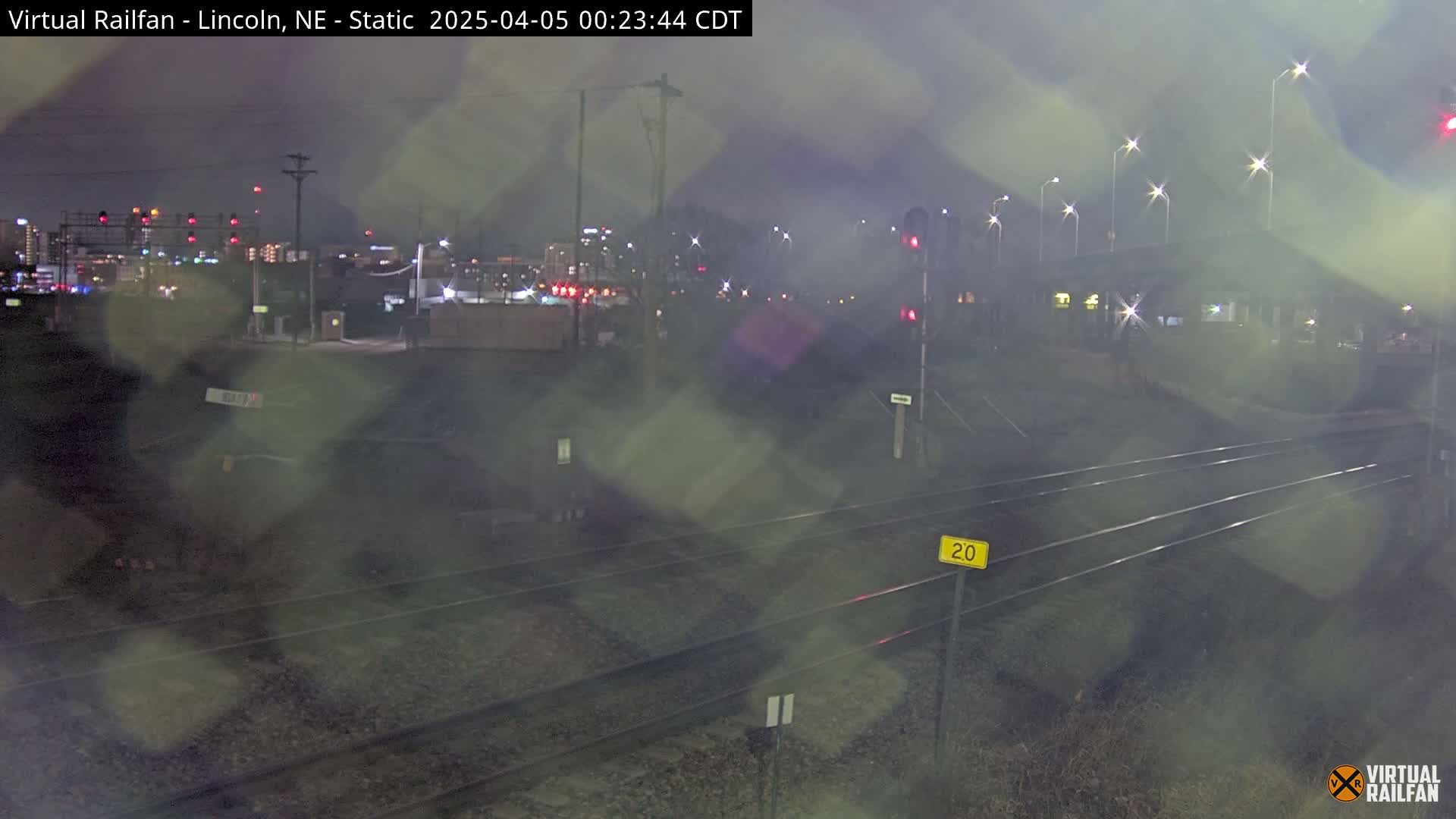 A nighttime view of rain-covered railroad tracks leading toward a city skyline under a dark, overcast sky.