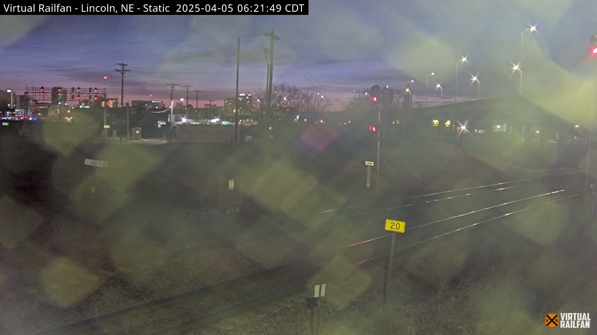 A twilight view of railroad tracks and a city skyline in the distance, under a partly cloudy sky.
