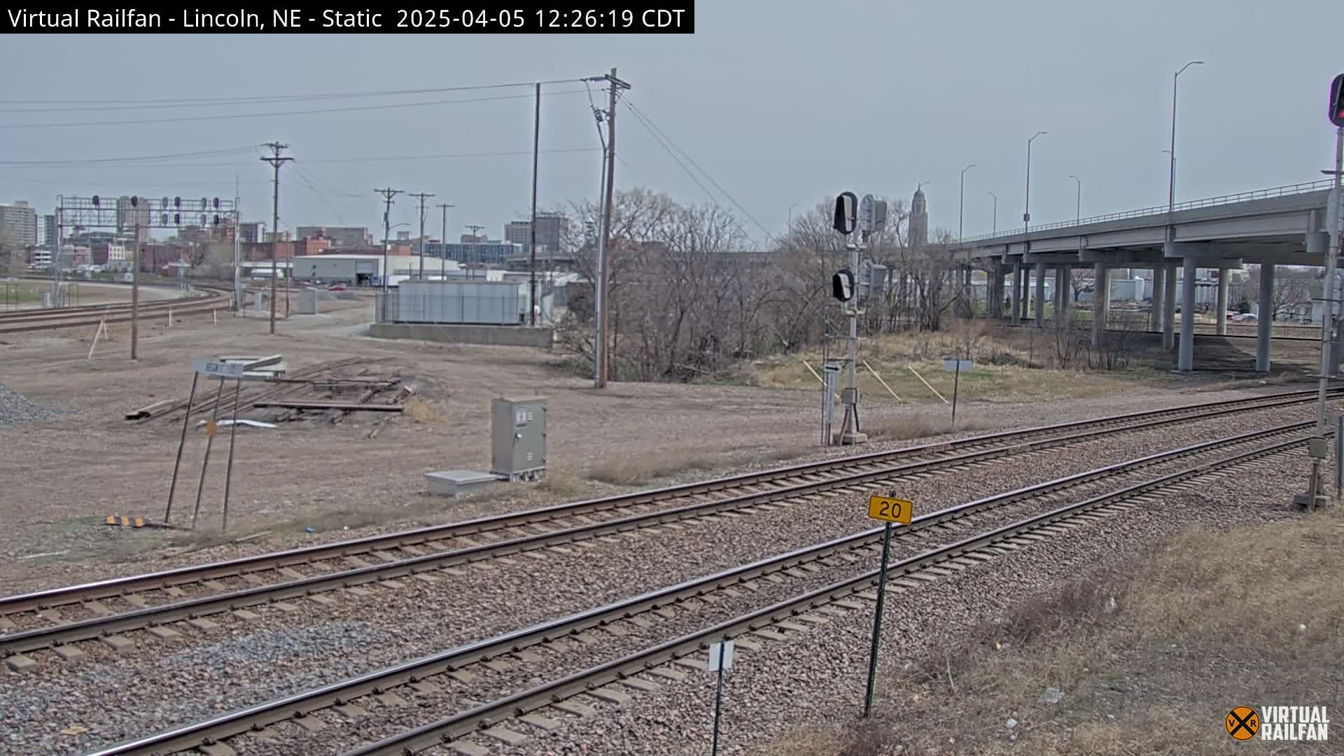 A view of railroad tracks under a highway overpass on a cloudy day, with bare trees and a city skyline in the background.