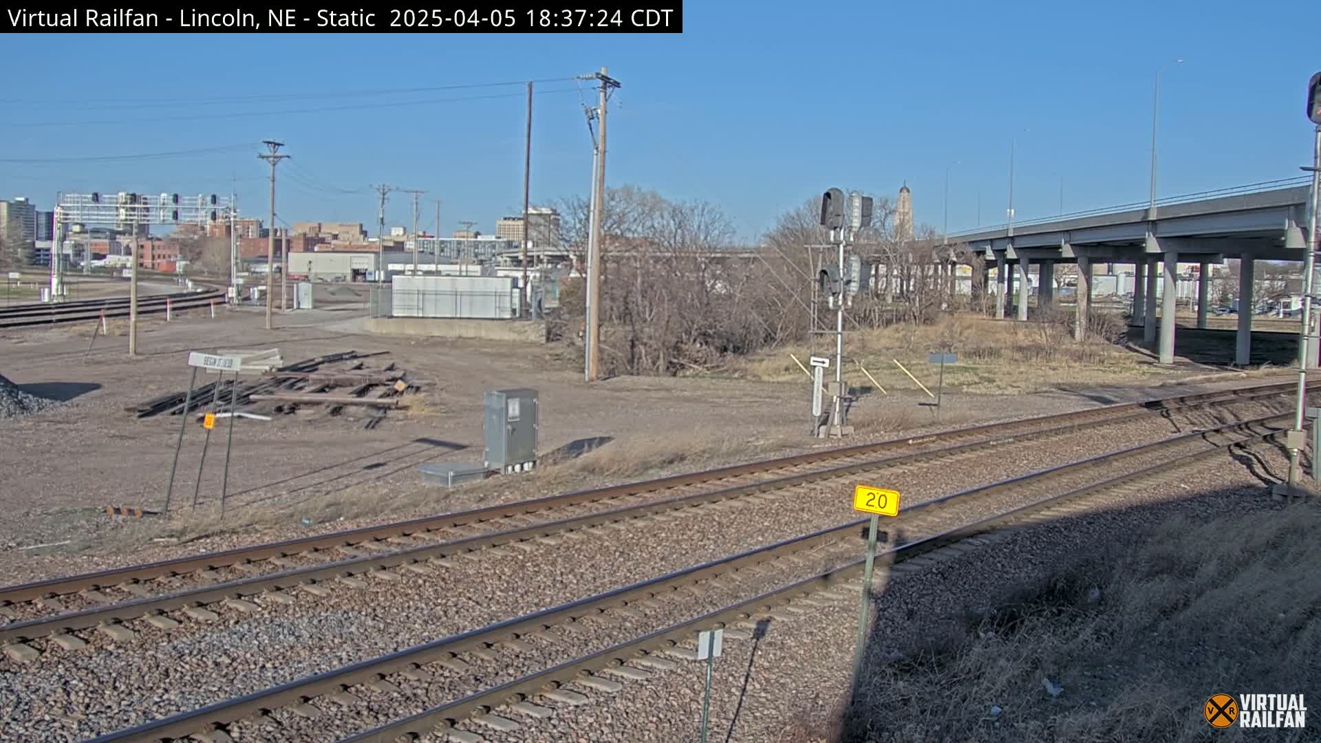 Here is a description of the image in a single sentence:  A sunny day reveals a section of railroad tracks near a city skyline, with a highway overpass in the background and some discarded materials in the foreground.