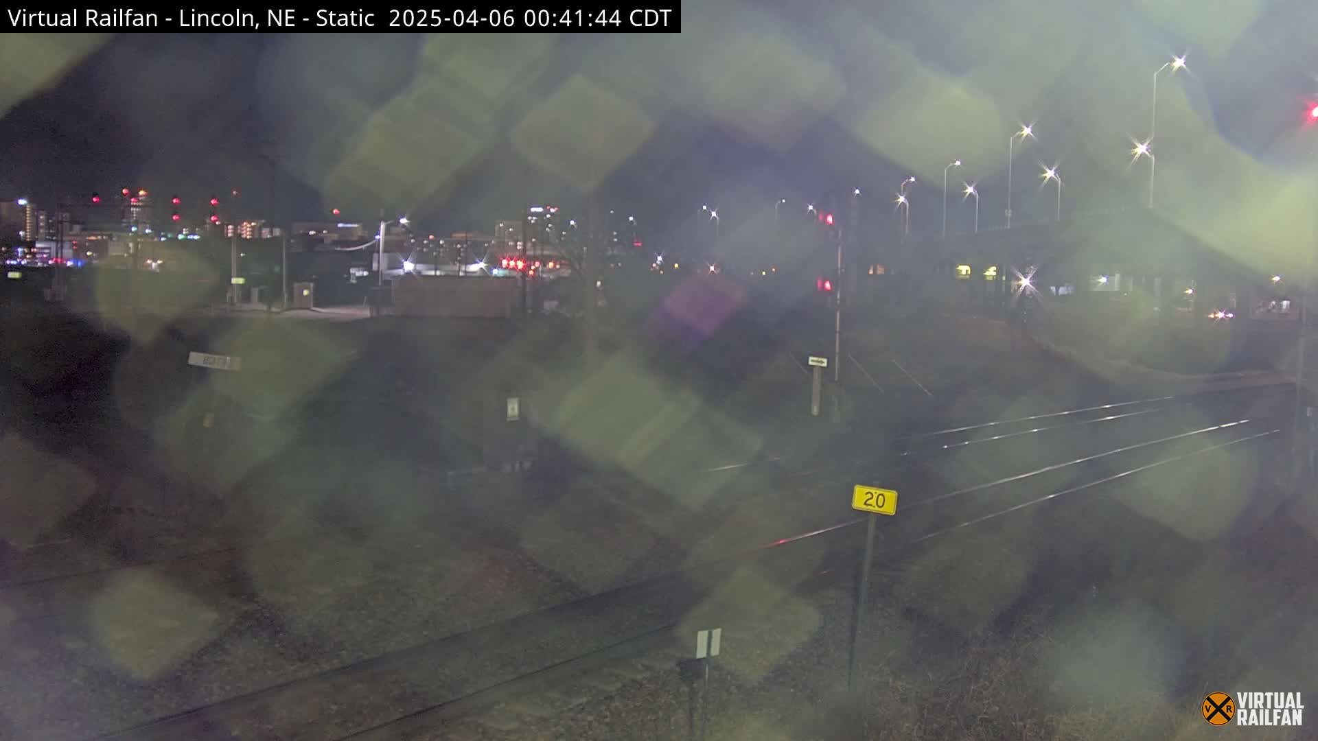 A nighttime view of railroad tracks and a city skyline under rainy conditions.