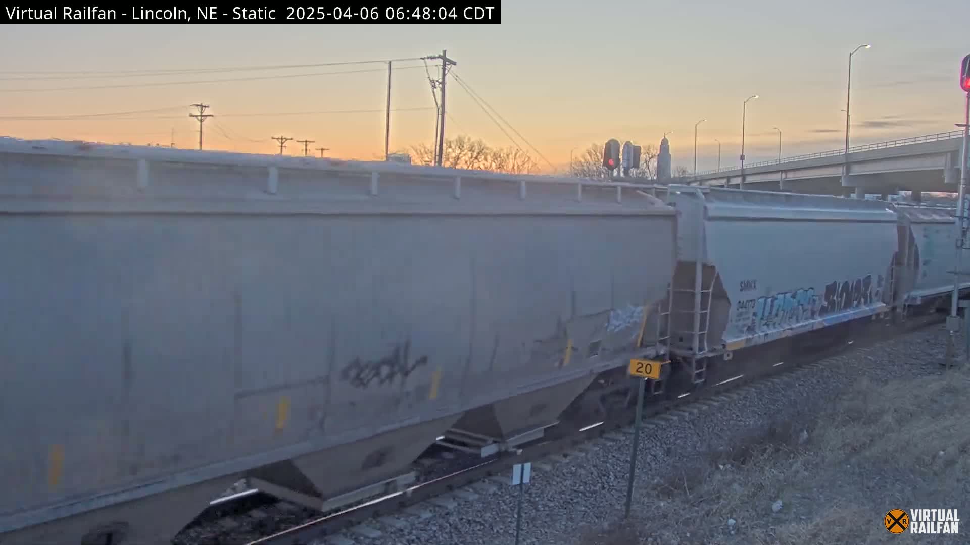 A long freight train with graffiti on some of its cars is moving past a road overpass at dawn.