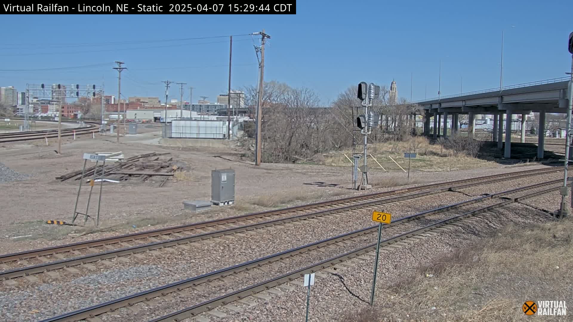 A view of railroad tracks on a clear, sunny day, with a city skyline in the background and a highway overpass visible.