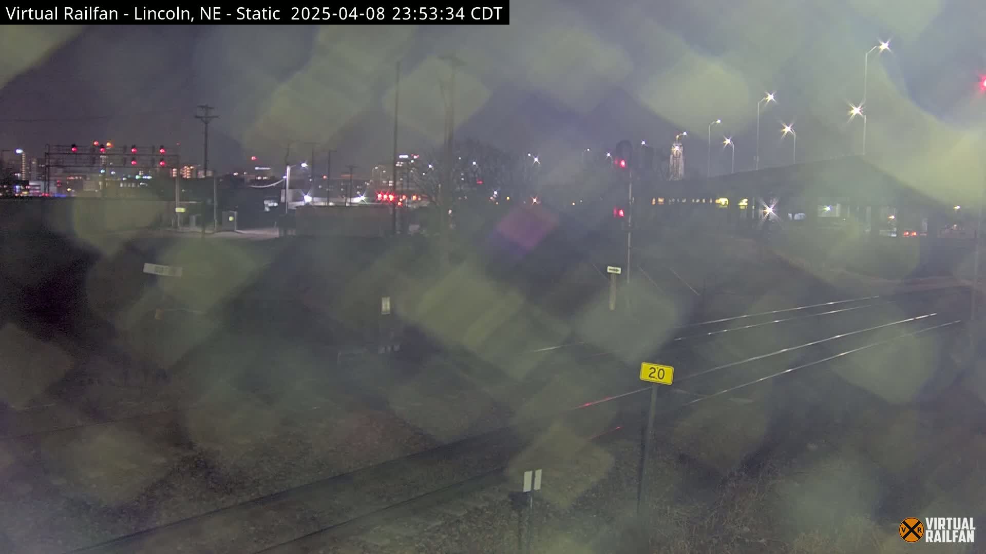 A nighttime view of railroad tracks and a city skyline in the distance, under slightly hazy conditions.