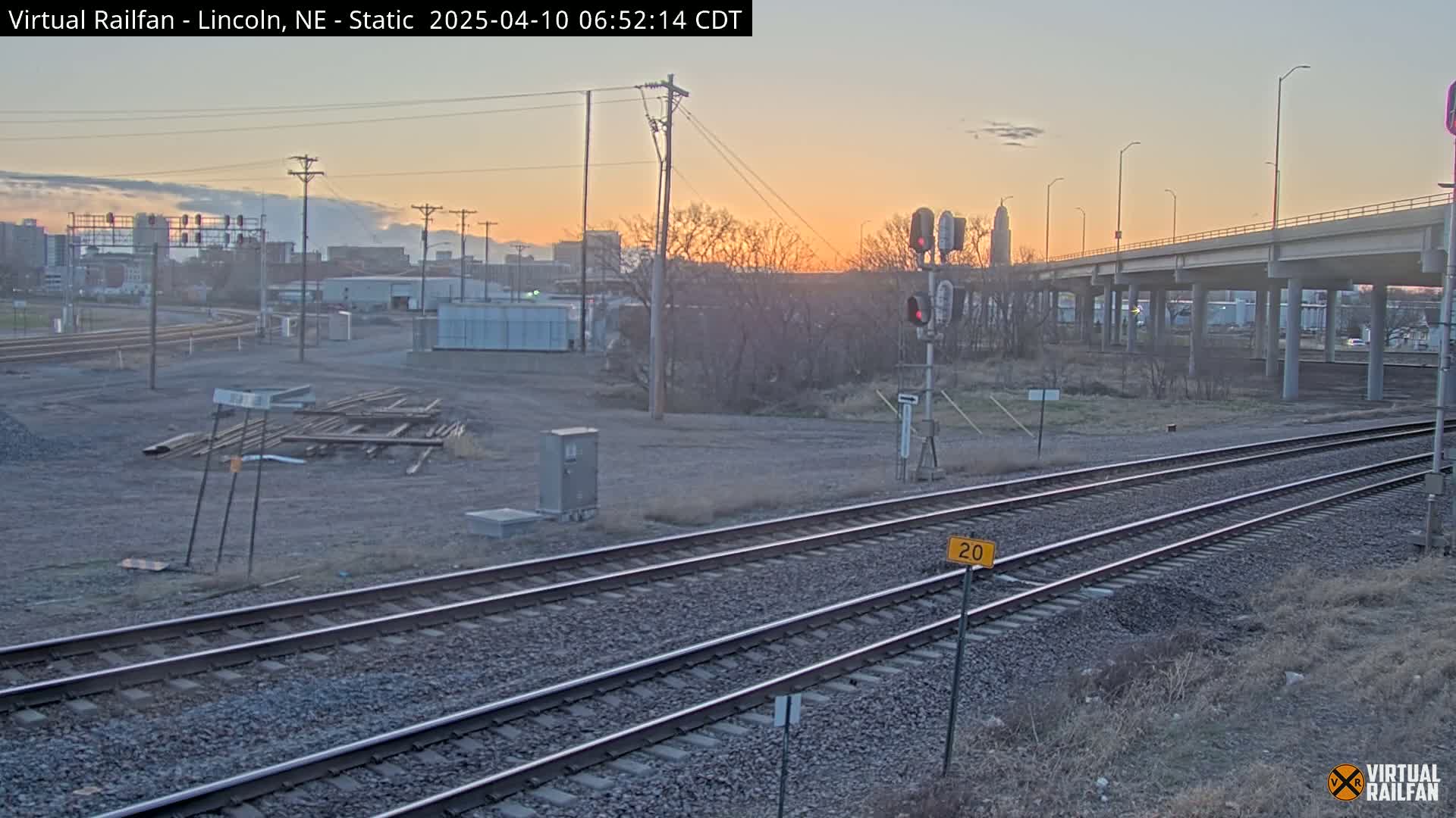 A view of railroad tracks at sunset, with a highway overpass in the background and some industrial buildings in the distance.