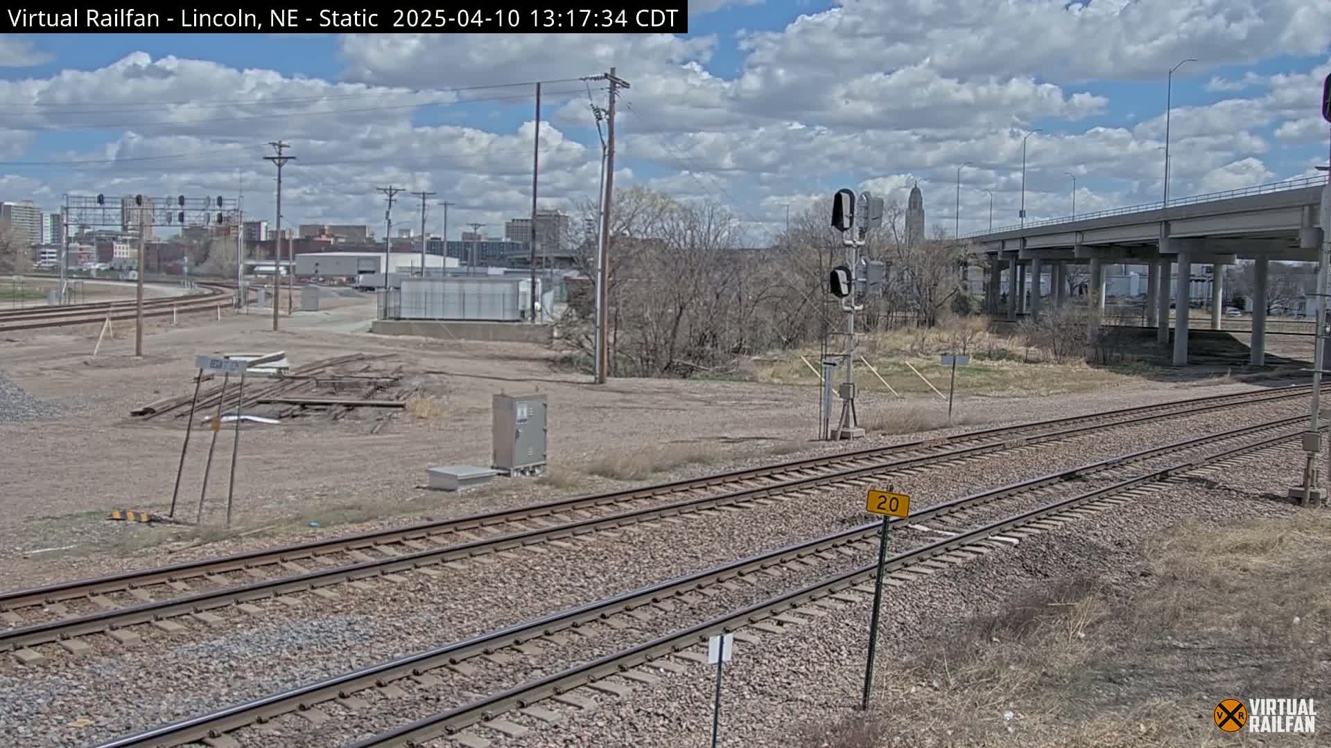 A view of railroad tracks under a partly cloudy sky, with a highway overpass in the background and a city skyline visible in the distance.