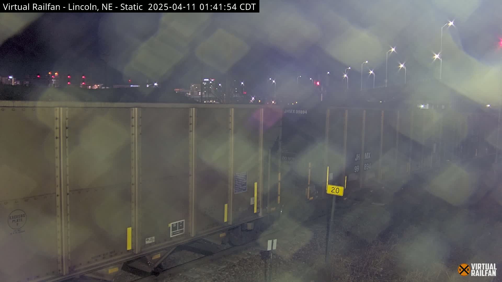 A nighttime view of a freight train car, partially obscured by rain or mist, with city lights visible in the background.