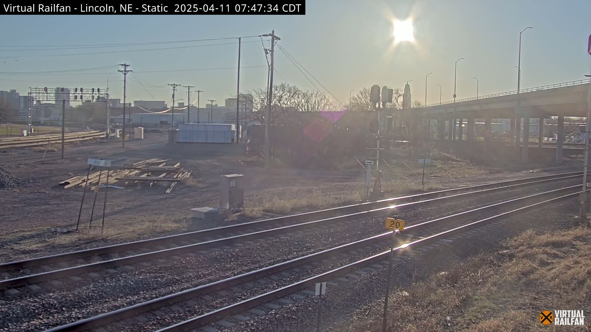 A sunny day reveals a railway yard with train tracks, some construction materials, and a highway overpass in the background.